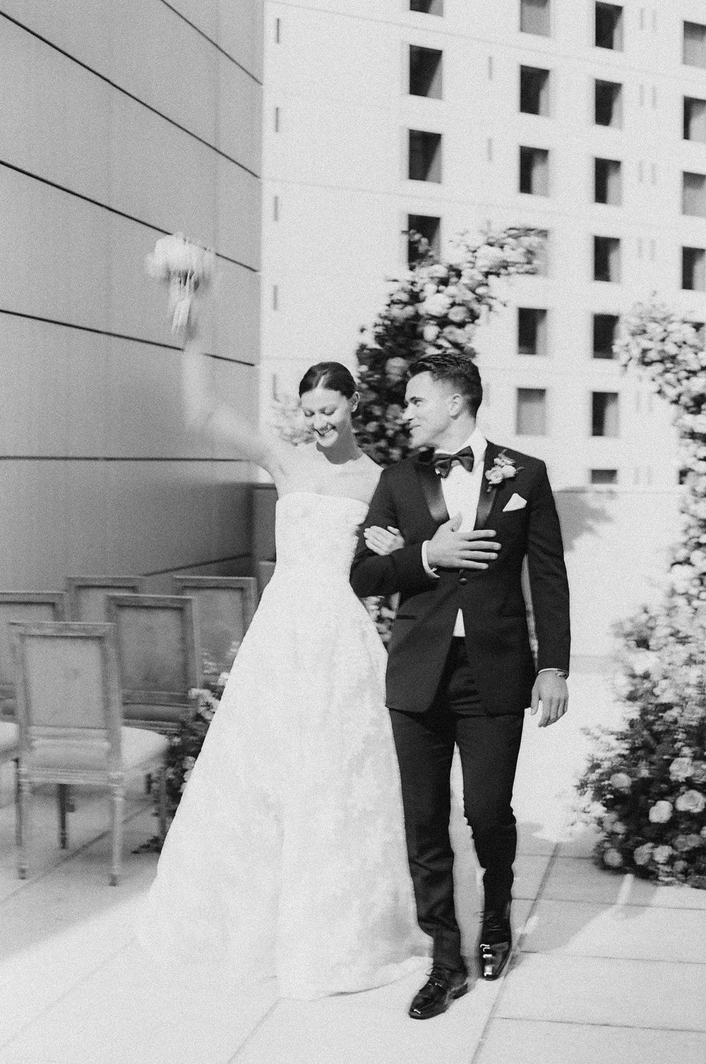 A couple in wedding attire walks arm in arm along a building exterior in classic black and white photography.
