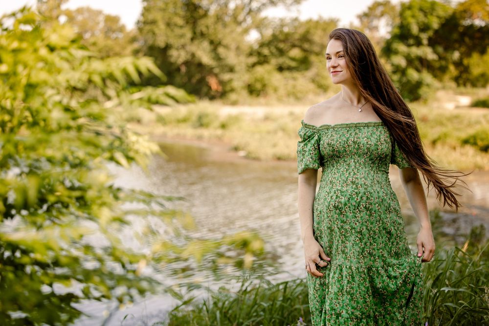 A figure in a flowing green off-shoulder dress stands gracefully near a scenic stream in a summer forest setting in fort worth