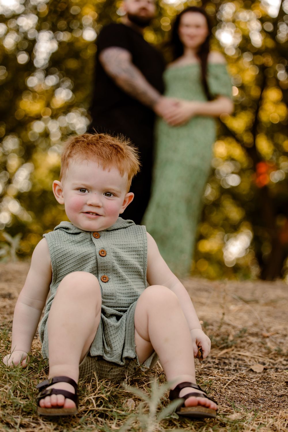 Young child in a gray sleeveless romper and sandals sits on ground with blurred figures in background on sunny day in fort worth