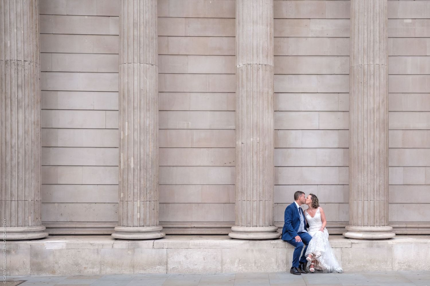 A couple walks down an elegant marble hallway on their wedding day, with ornate wooden doors in the background.