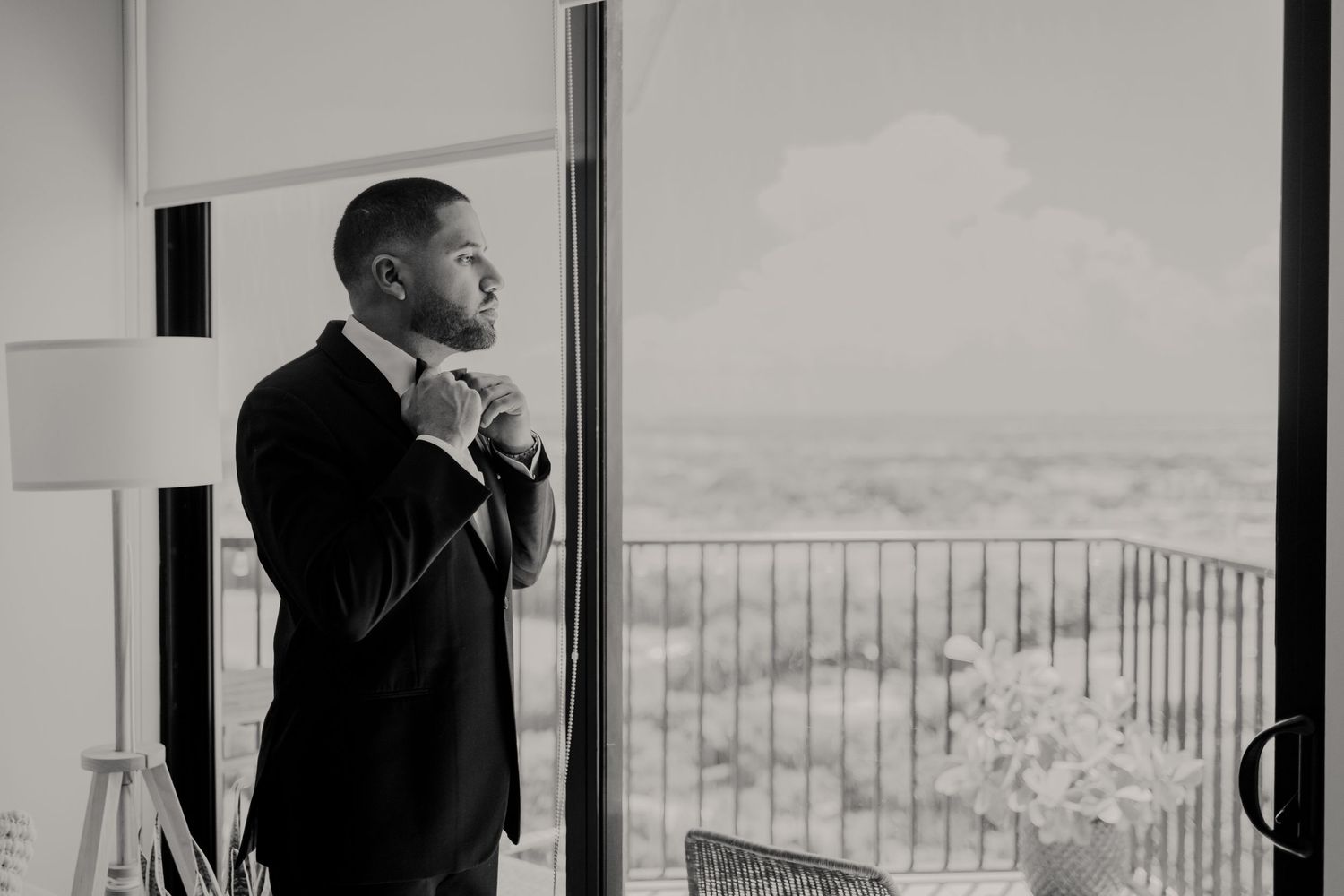 A groom in a dark suit adjusts his tie while looking thoughtfully out a window with a scenic view.