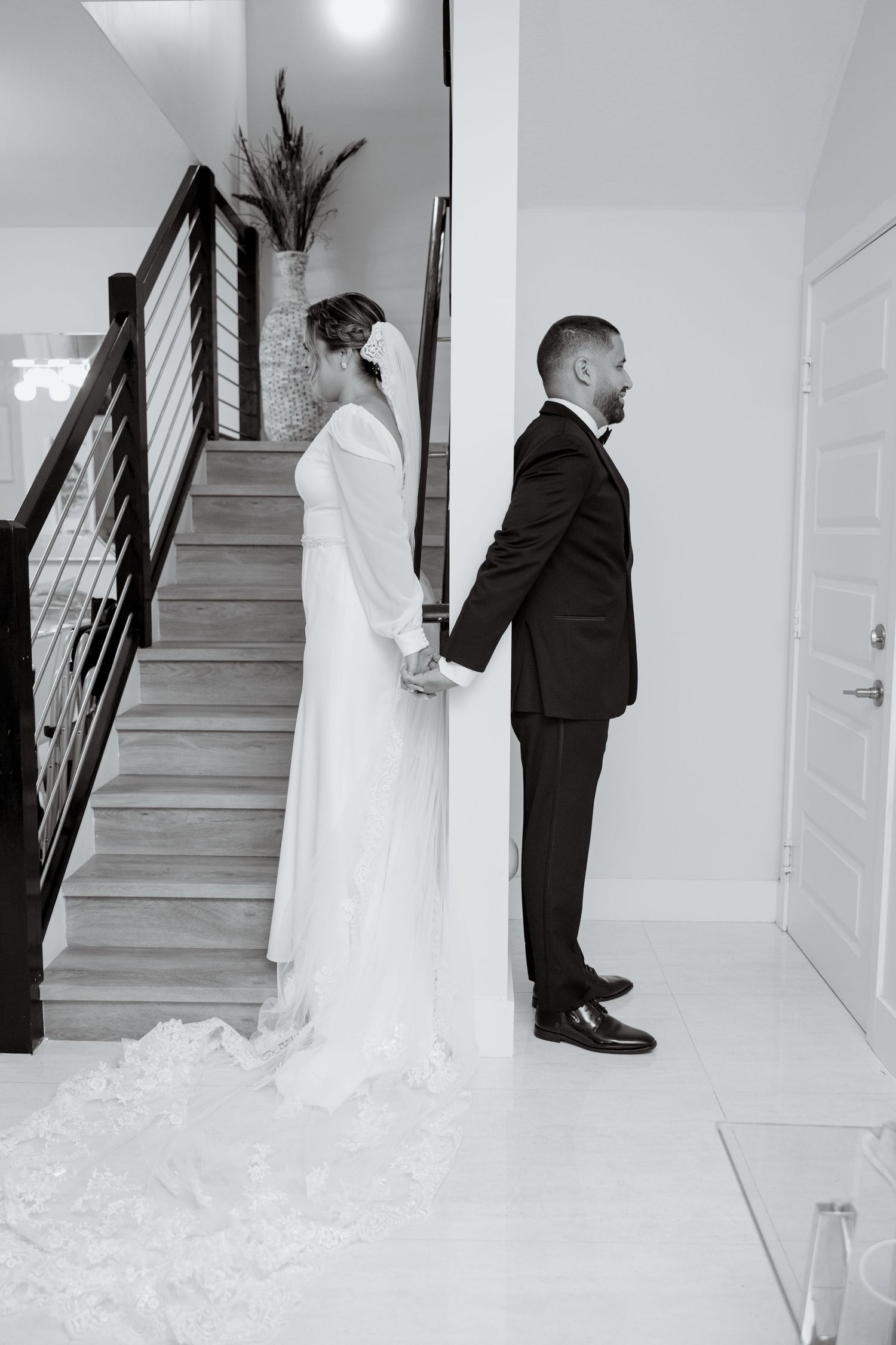 Black and white photo of a bride and groom holding hands while standing back to back on a staircase.
