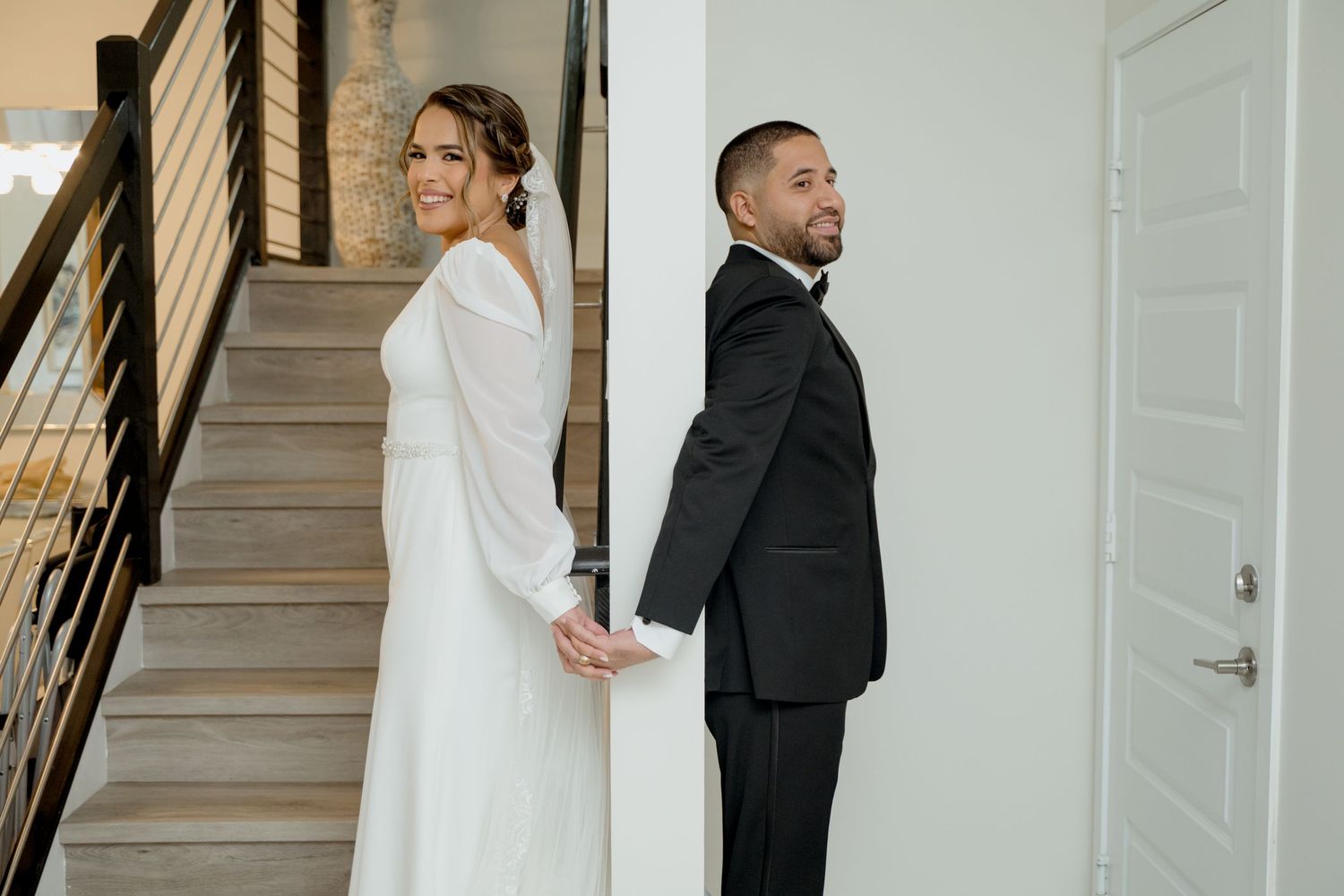 Bride and groom stand back to back holding hands on staircase during first look wedding photo moment.