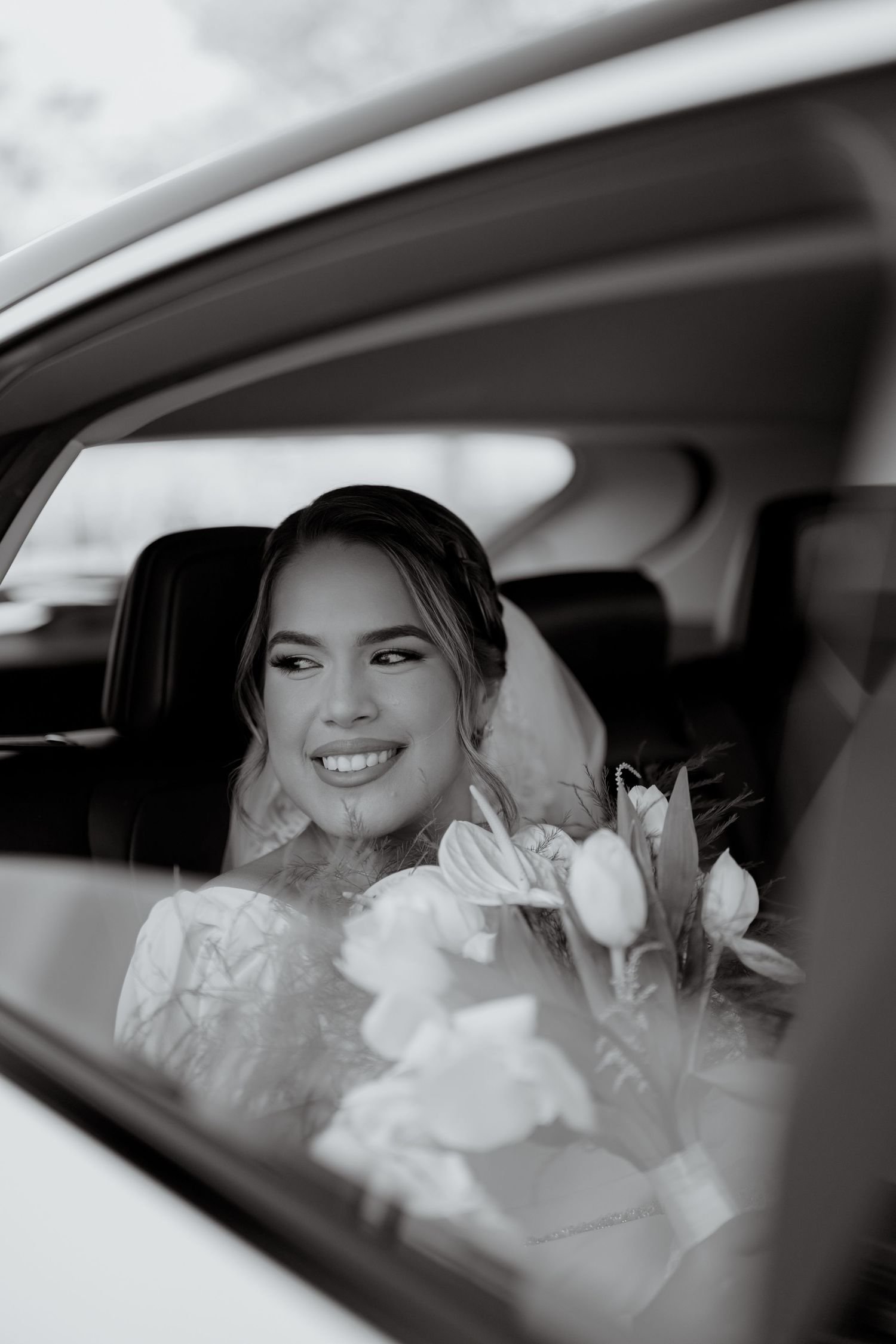 Black and white photo of a bride sitting in a car with a white flower bouquet and delicate lace wedding dress.
