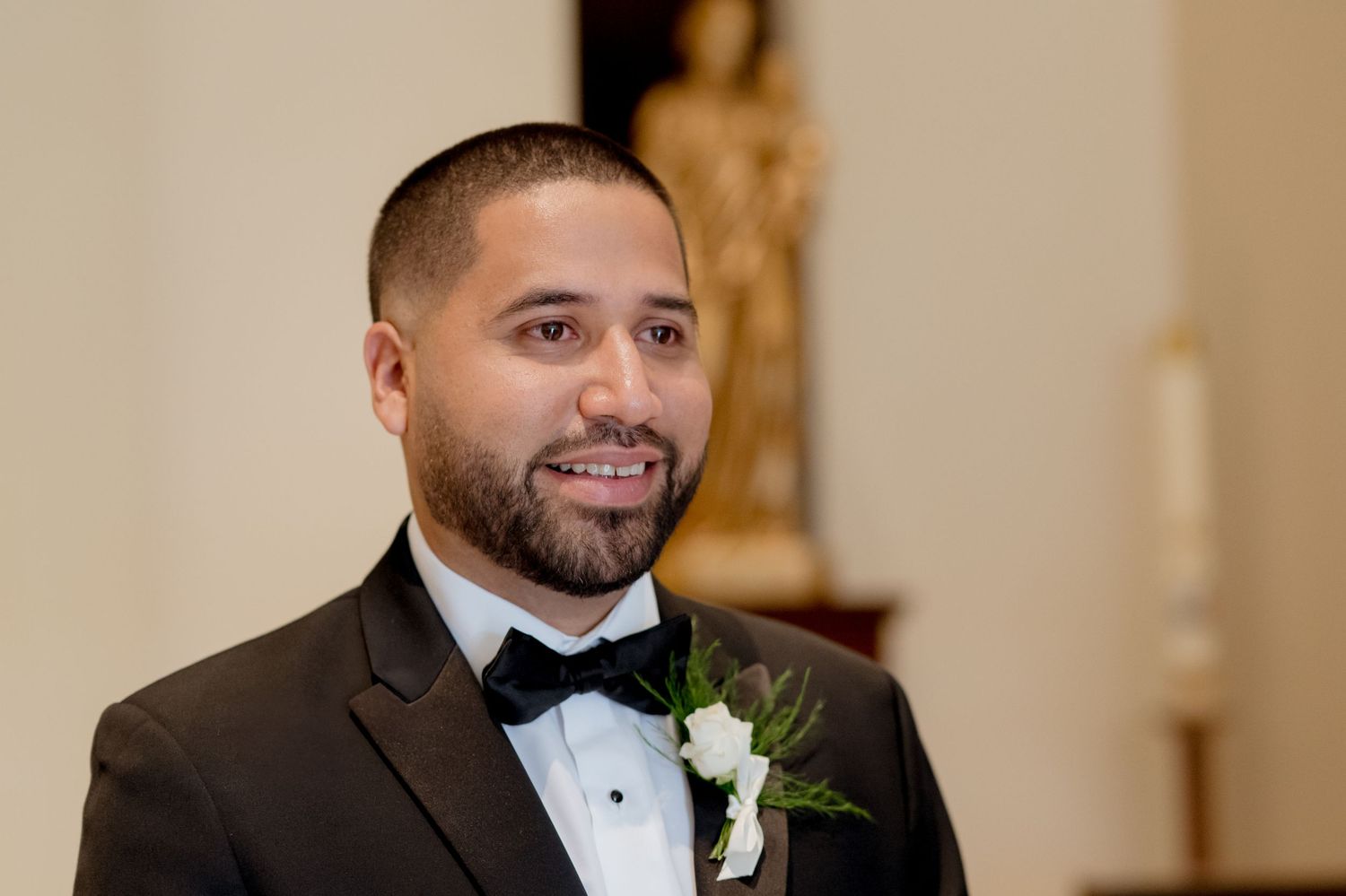 Man in black tuxedo and bowtie smiles warmly in a bright elegant indoor setting.