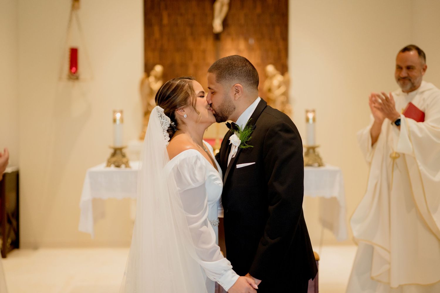 A couple shares a romantic kiss at the altar during their wedding ceremony while a priest looks on from behind.