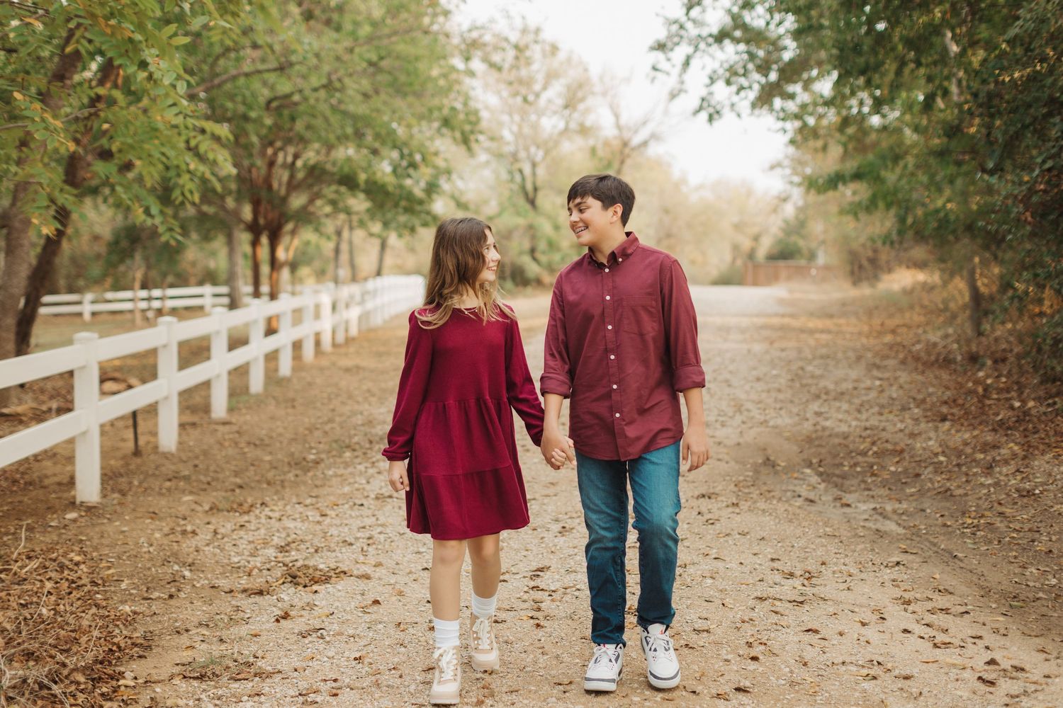 A couple in burgundy and denim walk hand in hand along a tree-lined path with white fencing in autumn.