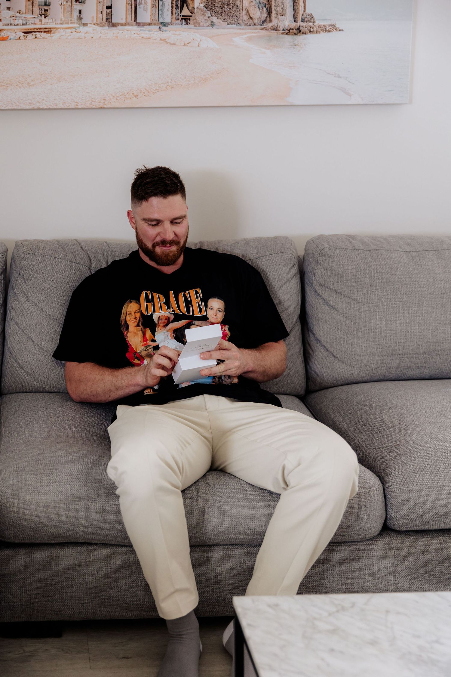 Someone relaxing on a gray couch using a mobile device while wearing a black t-shirt and white pants.