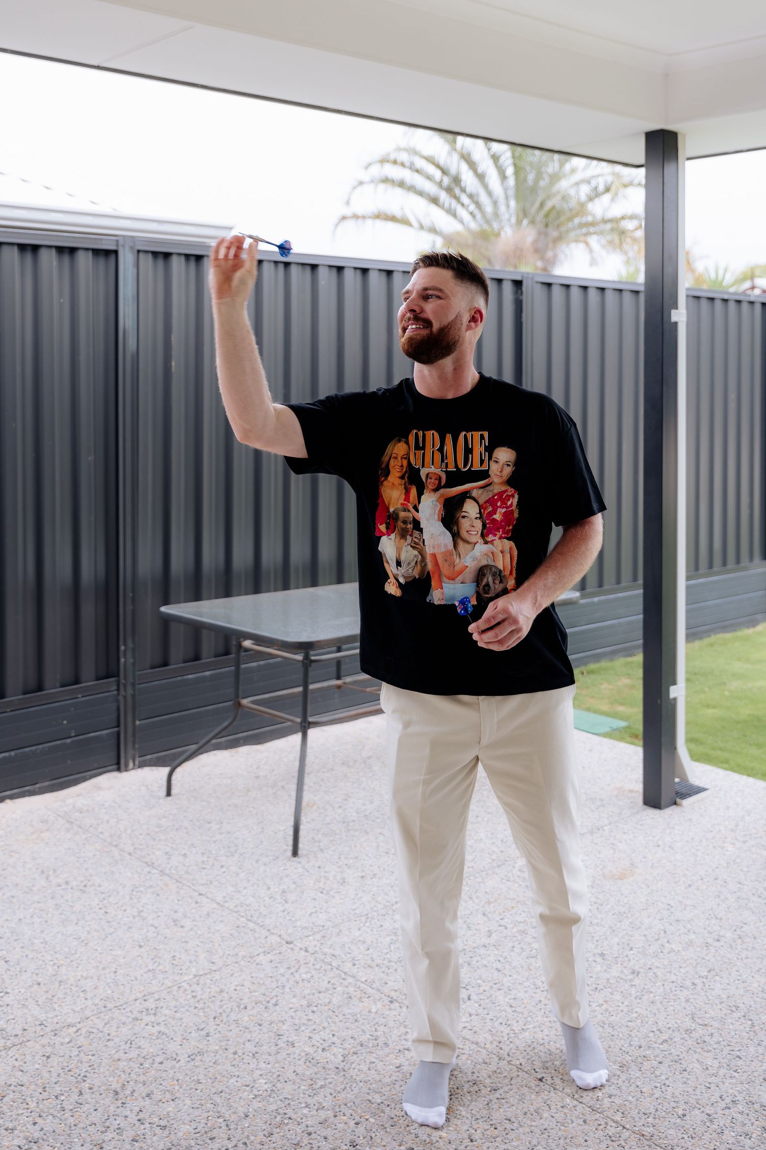 Someone wearing a casual black graphic t-shirt and white pants poses playfully outdoors on a patio with grey fencing.