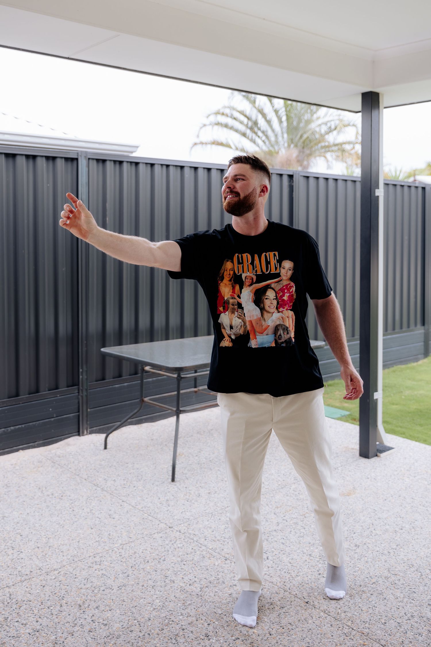 Person wearing a black graphic t-shirt and white pants poses casually on an outdoor patio with gray fencing.