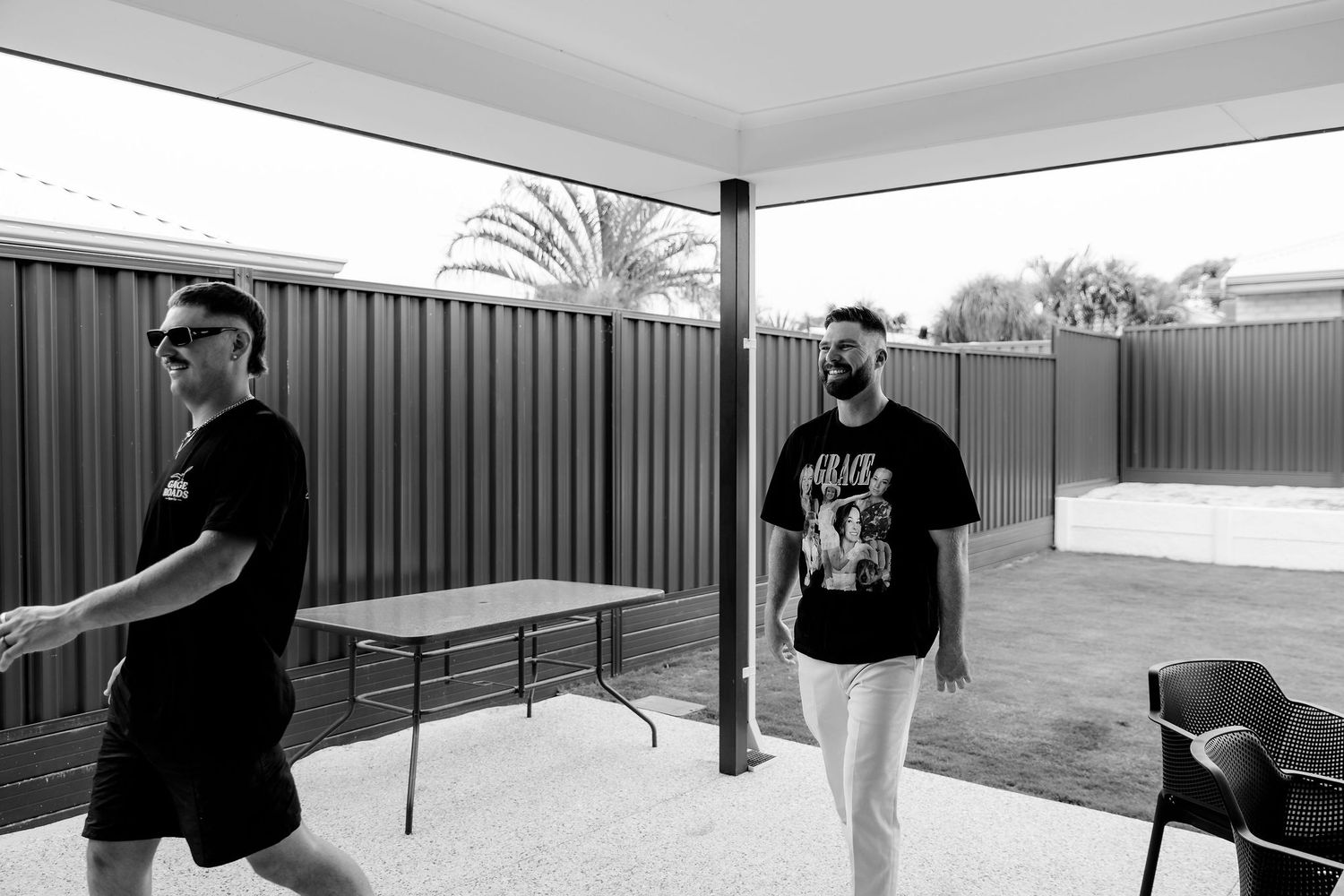 Two people wearing black t-shirts stand in an outdoor covered patio area with tables and chairs.