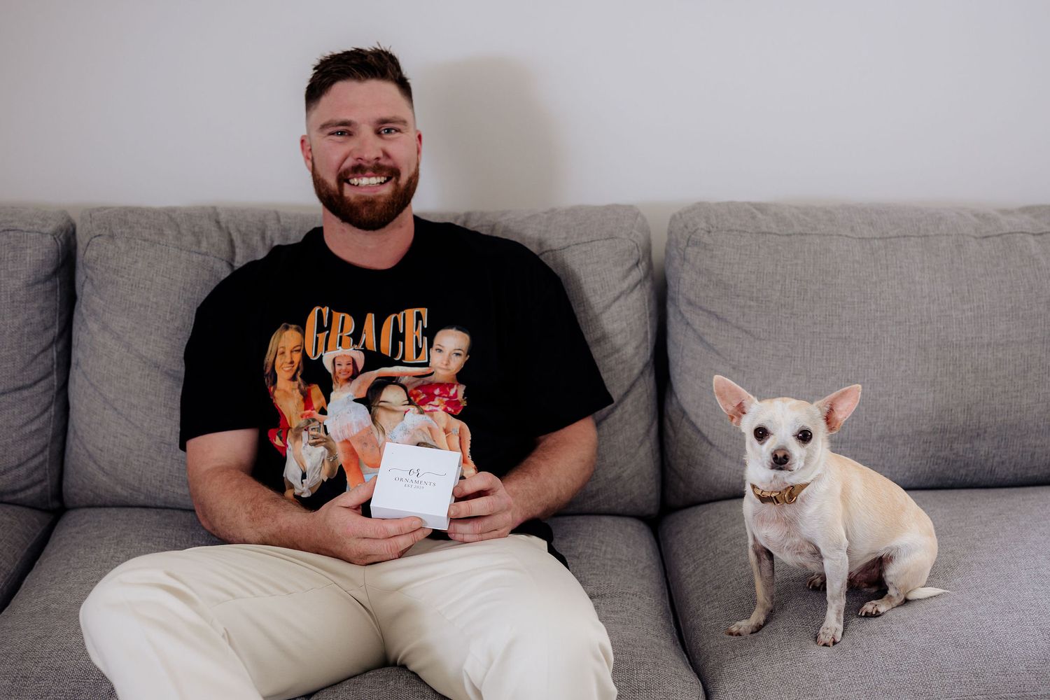 A small white chihuahua sits on a gray couch next to someone holding a white package.