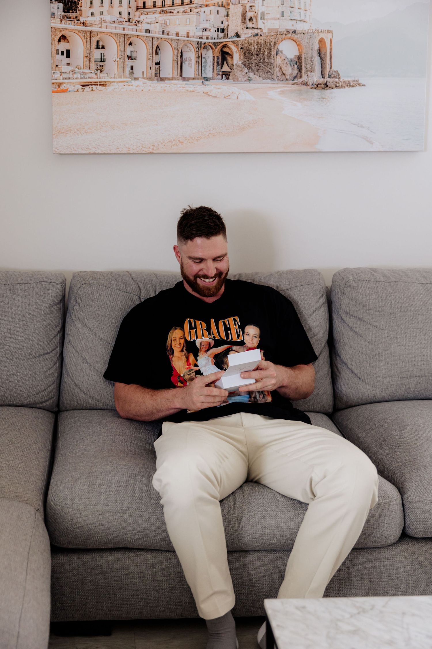 A person in a black graphic t-shirt and white pants sits on a gray couch looking down at a baby in their arms.