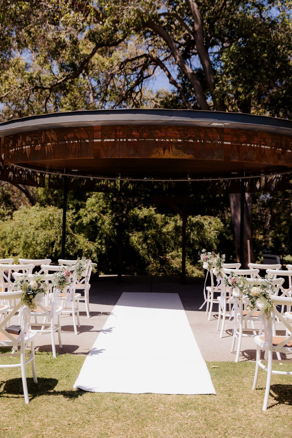 Outdoor wedding ceremony setup with white chairs and aisle runner under a round wooden pergola surrounded by trees.