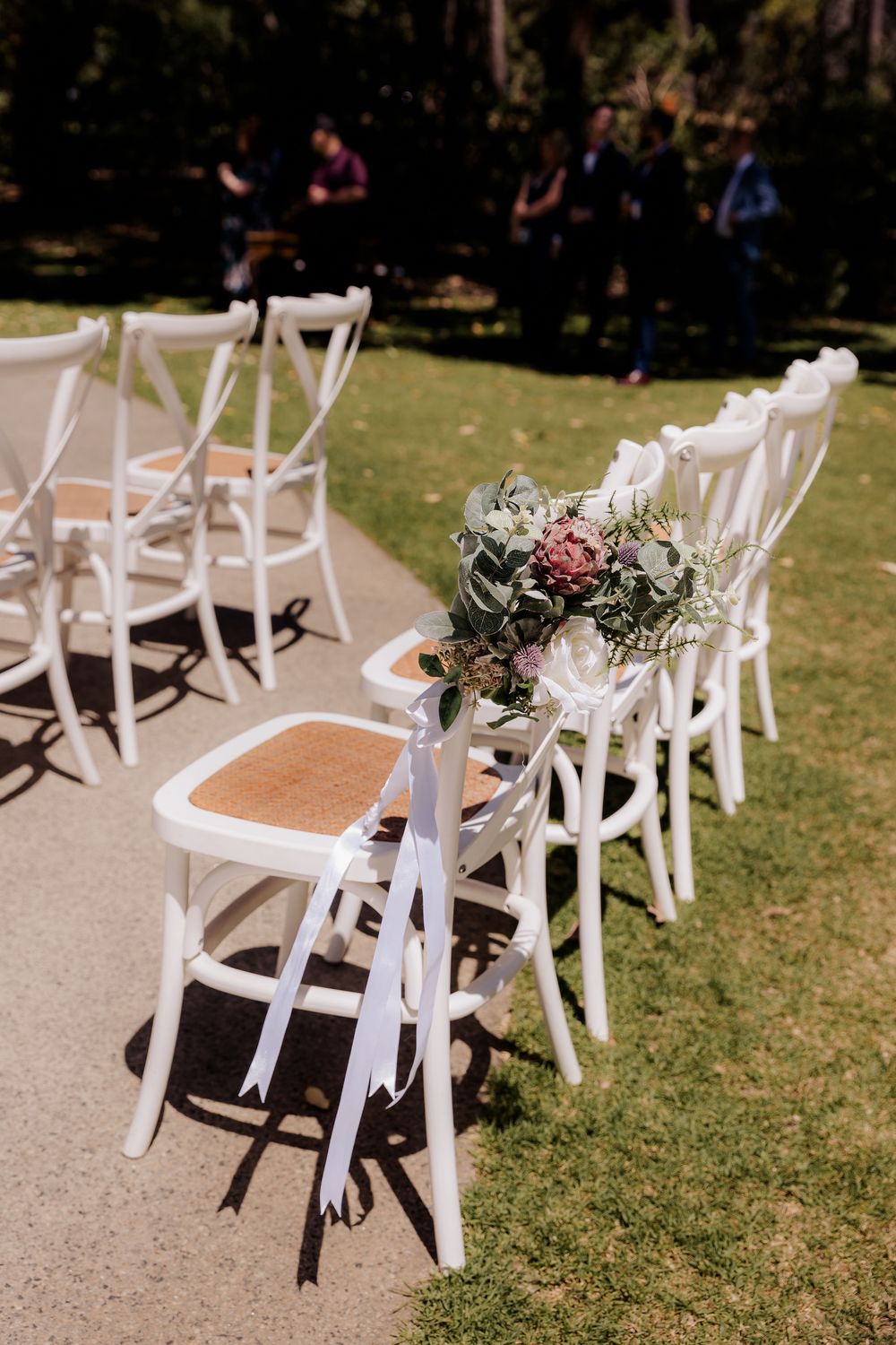 White crossback chairs lined up for an outdoor wedding ceremony with floral aisle decorations on a sunny day.