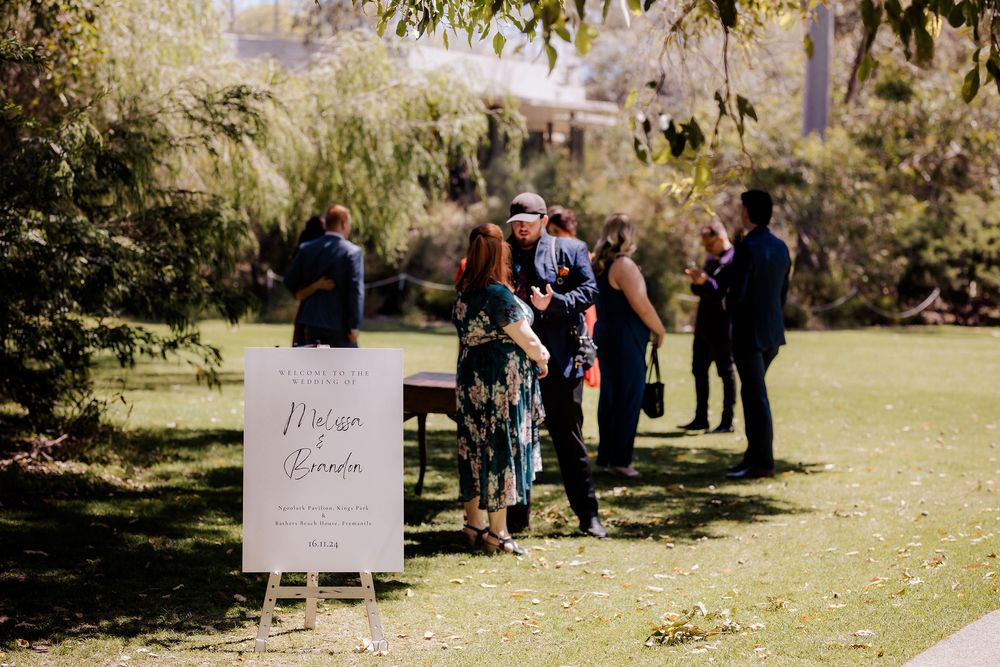Guests gather near a welcome sign at an outdoor wedding ceremony in a sunny garden setting.