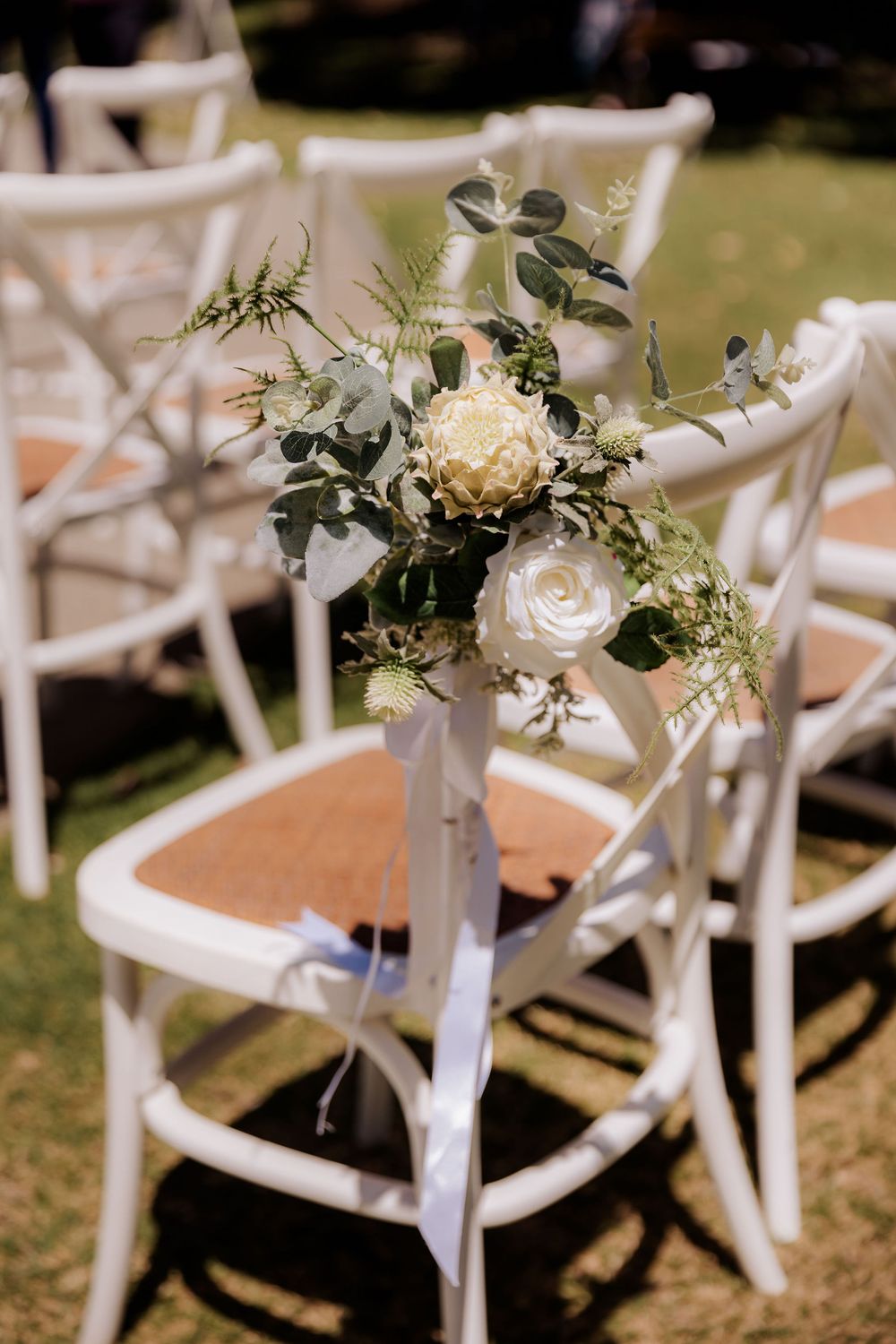 White floral arrangement with roses and eucalyptus decorates a crossback chair at an outdoor wedding ceremony setup.