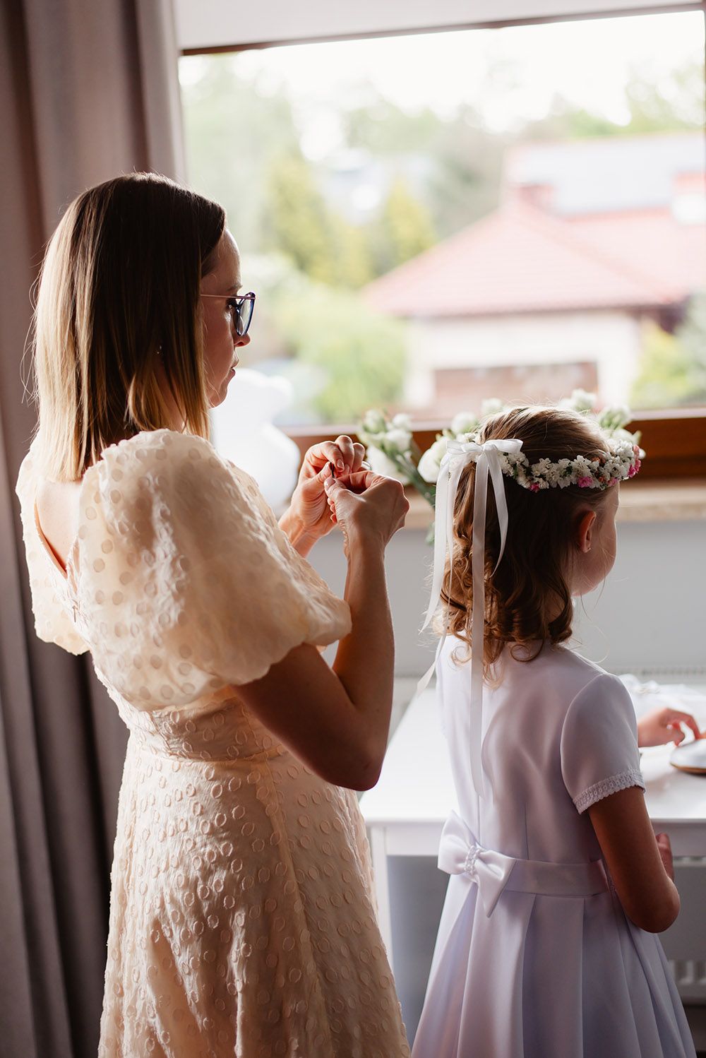 Mama poprawia wianek z białych kwiatów na głowie córki, przygotowującej się do Pierwszej Komunii Świętej, w tle jasne, naturalne światło.