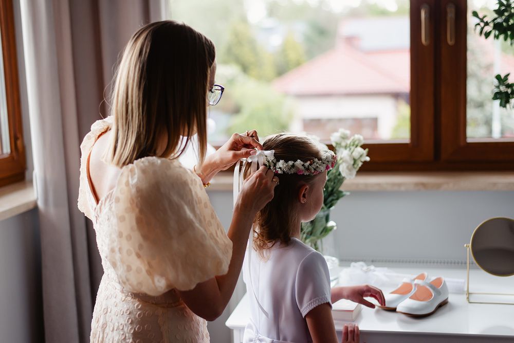 Mama poprawia wianek z białych kwiatów na głowie córki, przygotowującej się do Pierwszej Komunii Świętej, w tle jasne, naturalne światło.
