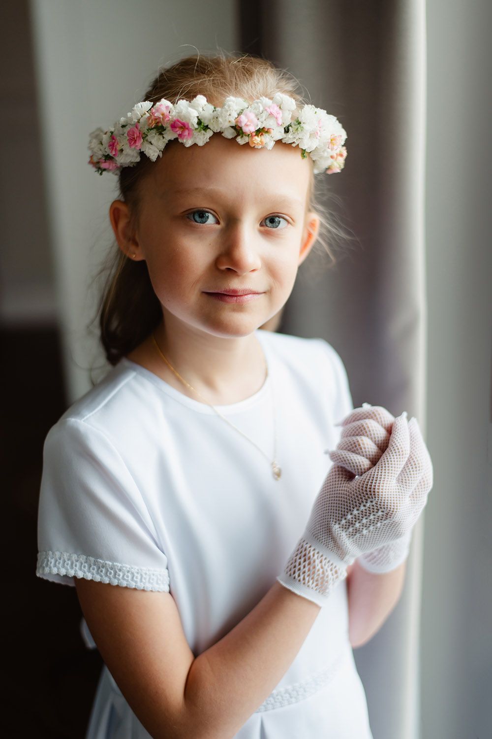 Portret dziewczynki w białej sukience komunijnej i wianku z białych kwiatów, sfotografowanej w miękkim, naturalnym świetle.