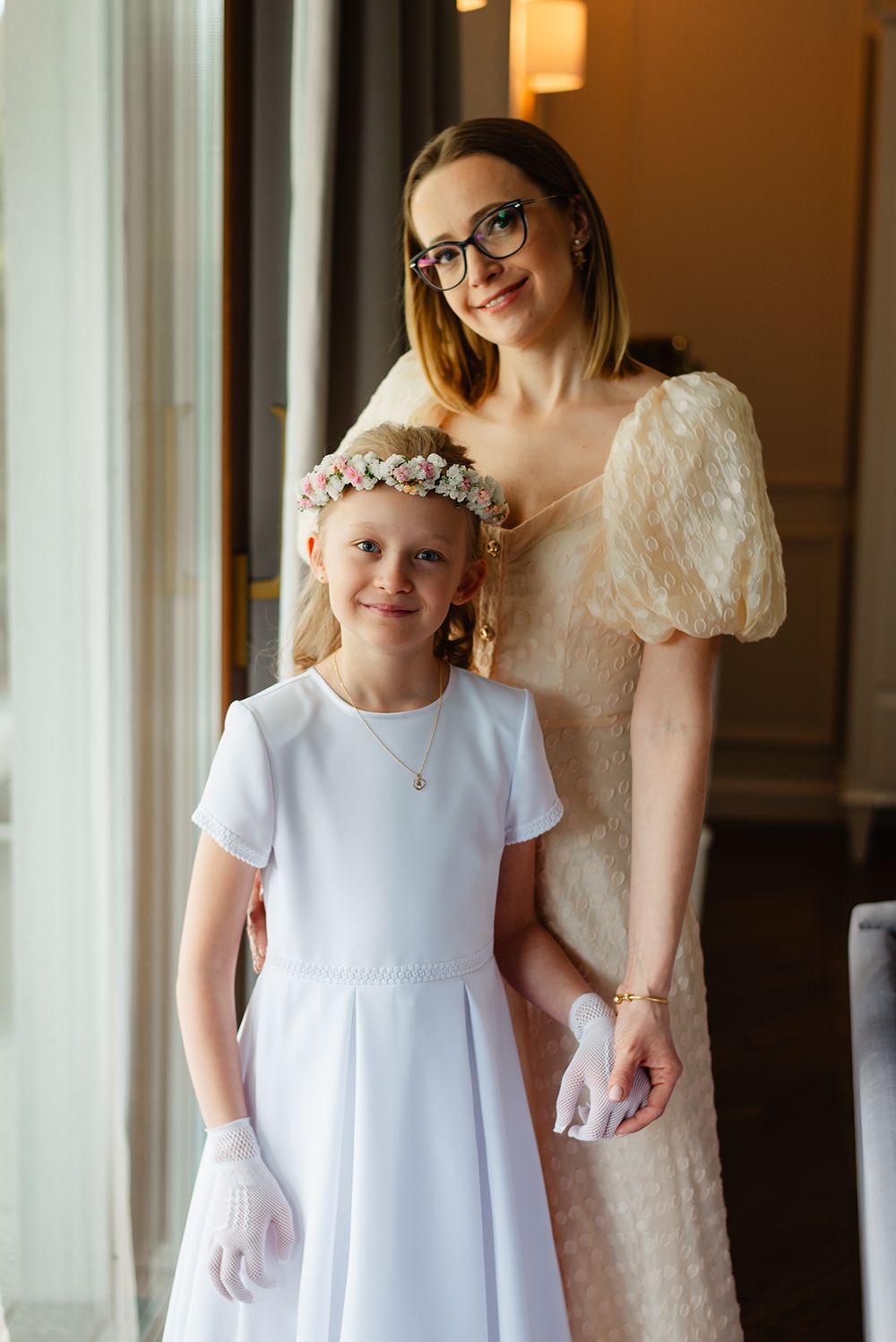 Portret komunijny – dziewczynka w wianku i mama w eleganckiej sukience stoją razem, patrząc z uśmiechem w obiektyw aparatu fotograficznego