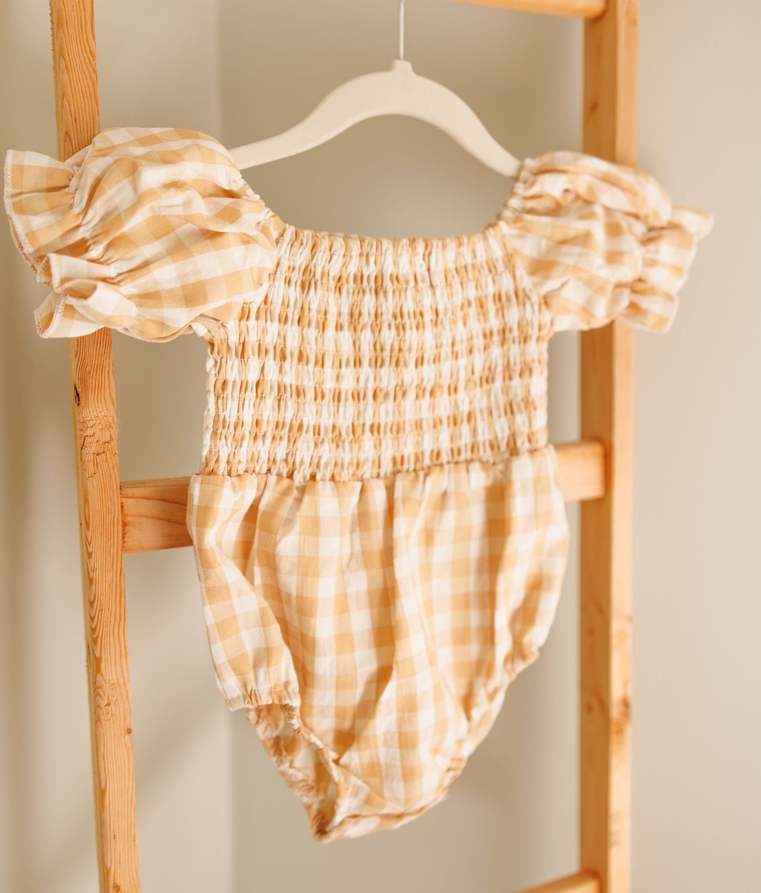 A peach gingham smocked romper with puff sleeves hanging on a white hanger against a wooden ladder.