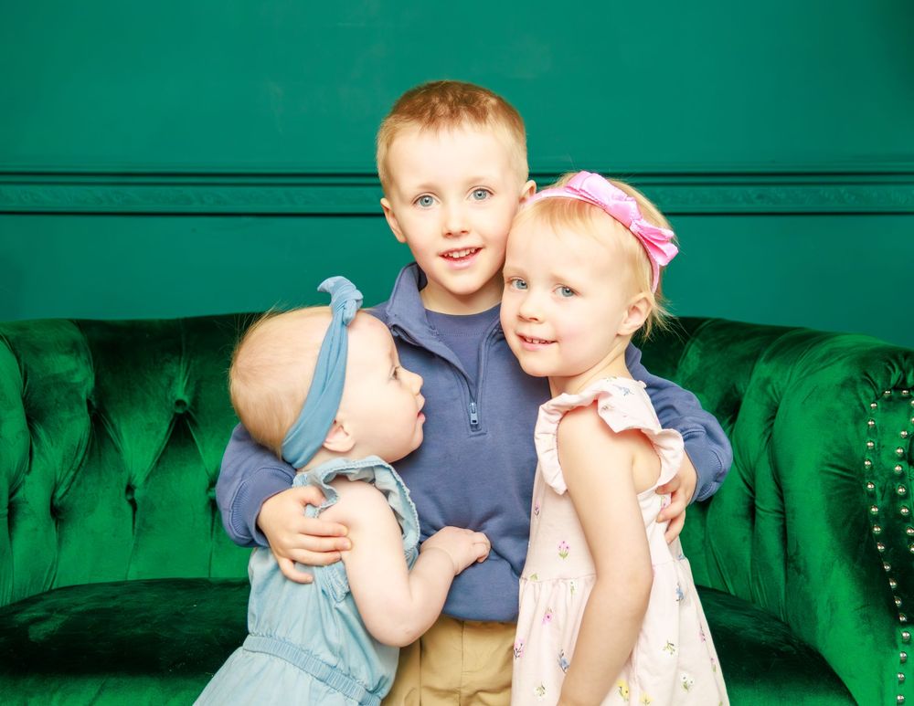 A bright studio portrait of three young children sitting together on an emerald green velvet couch.