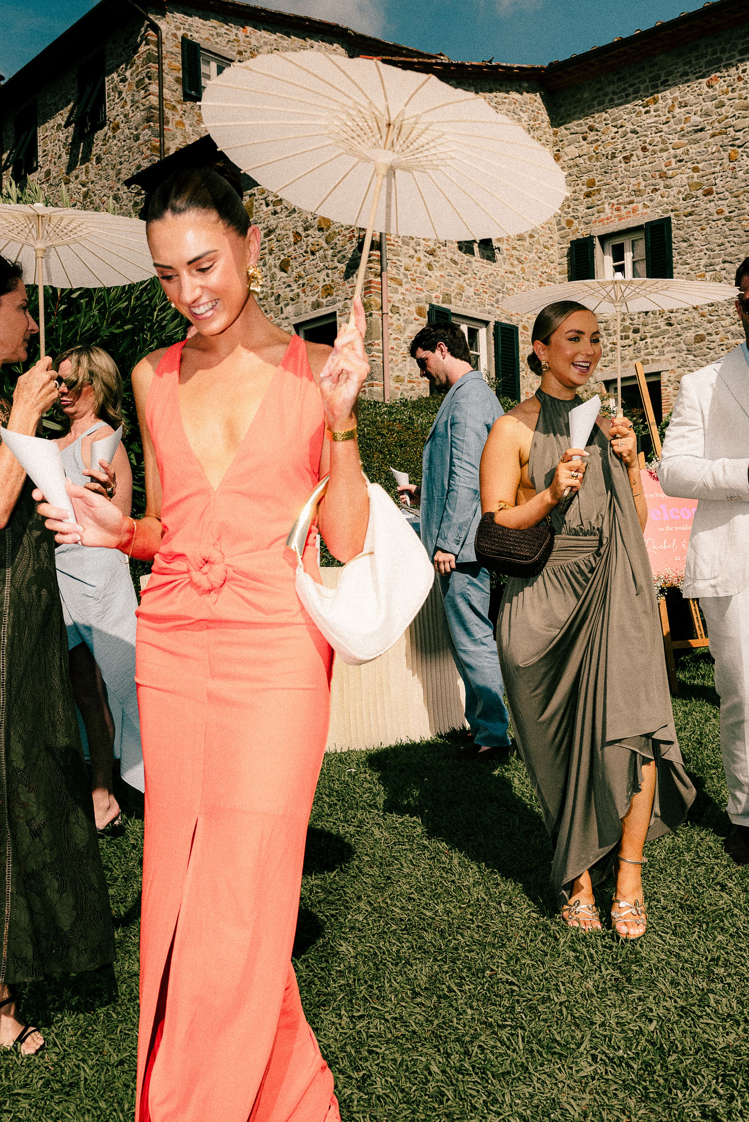 Elegantly dressed guests with parasols gathered on a lawn outside a rustic stone building during a summer event.