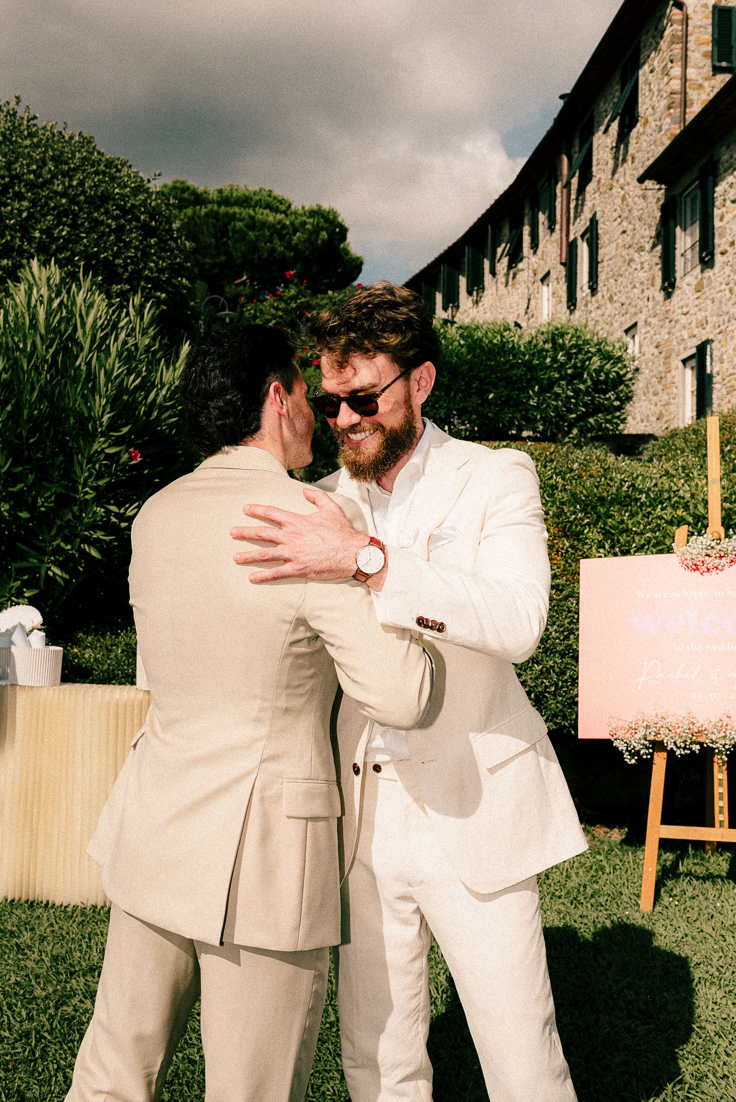Two people wearing light colored suits share an emotional embrace during an outdoor celebration at a rustic venue.