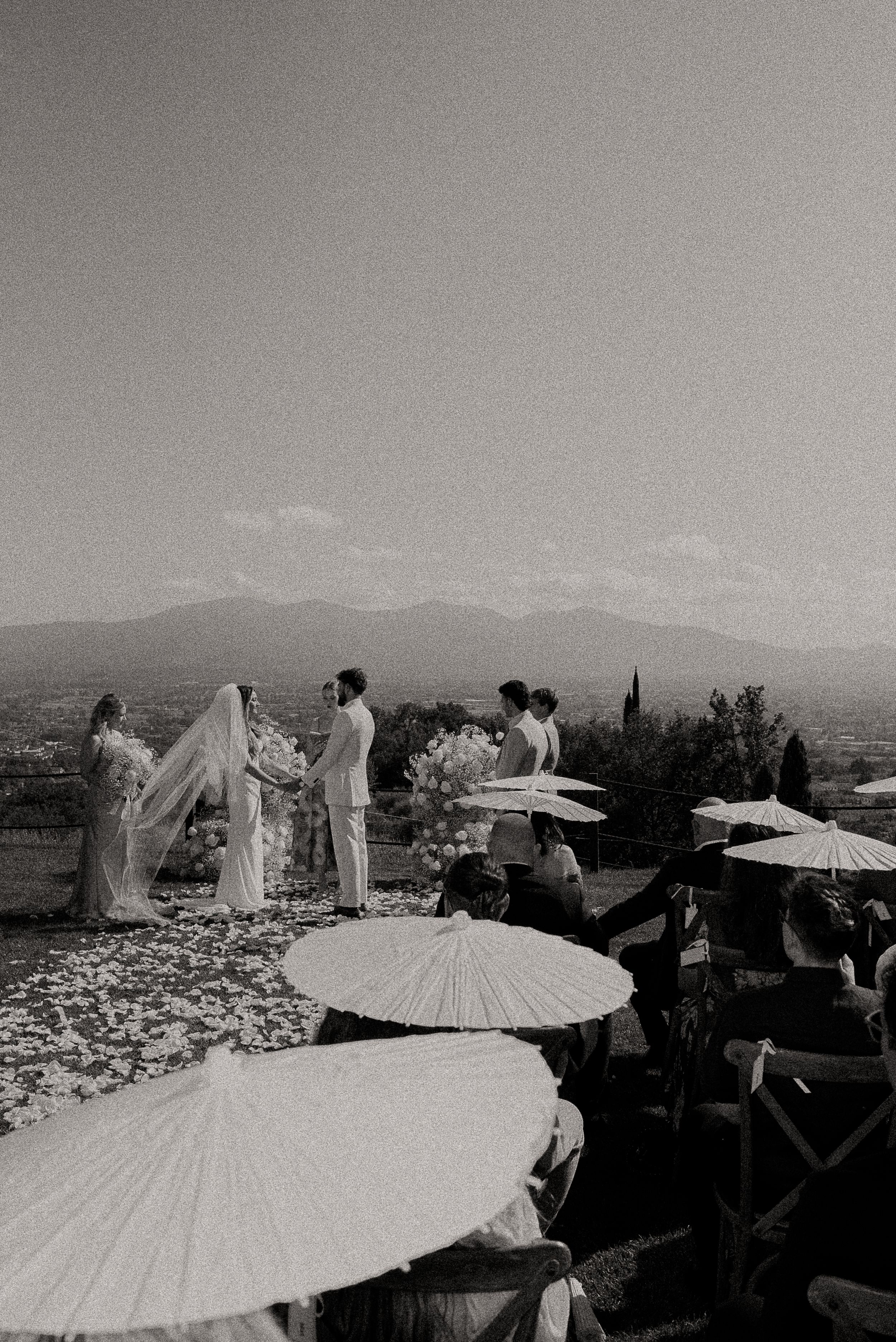 Black and white wedding scene with white parasols against mountain backdrop.
