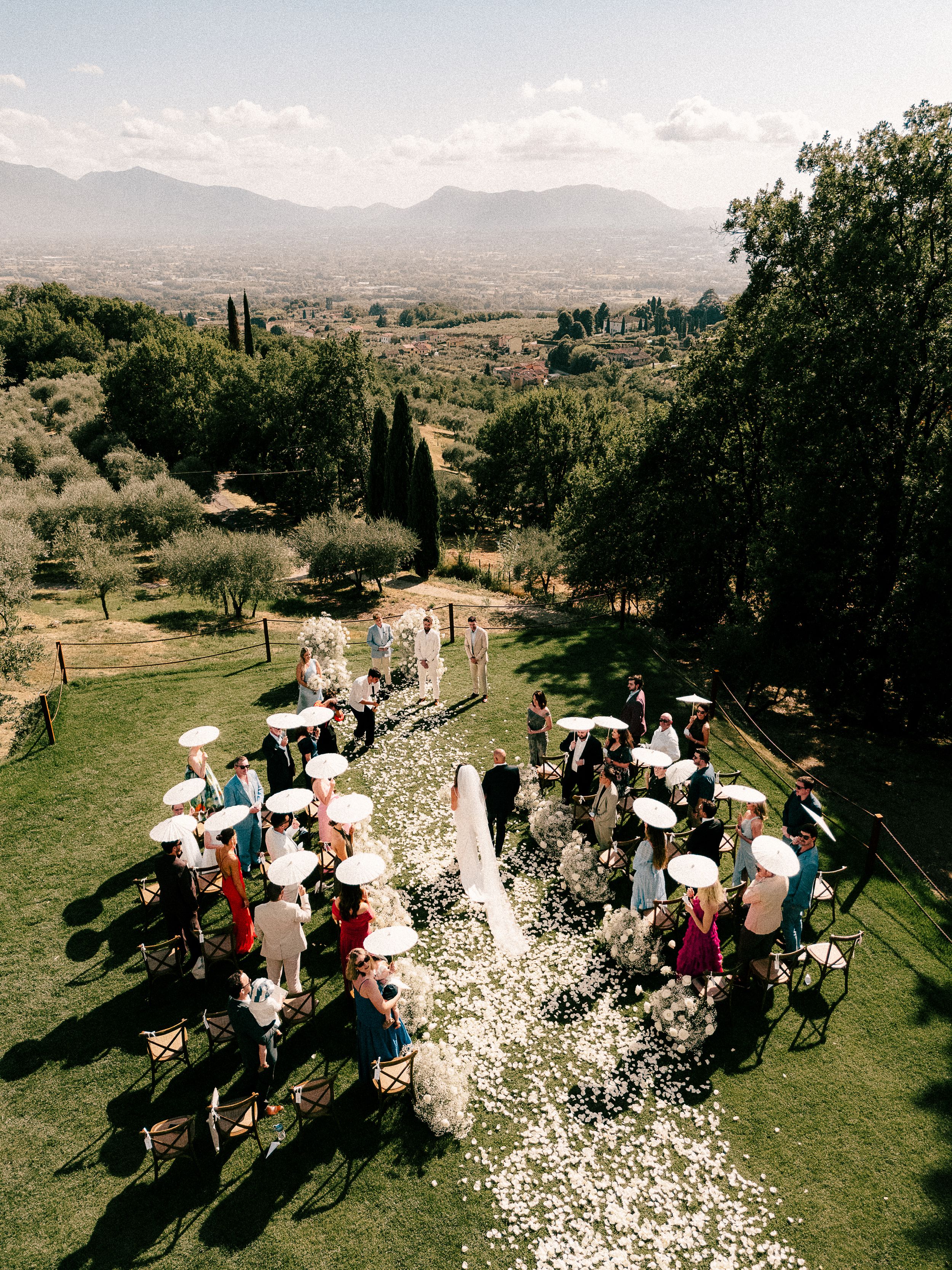 Outdoor wedding ceremony seating on a grassy hillside with mountains in the background and guests holding parasols.