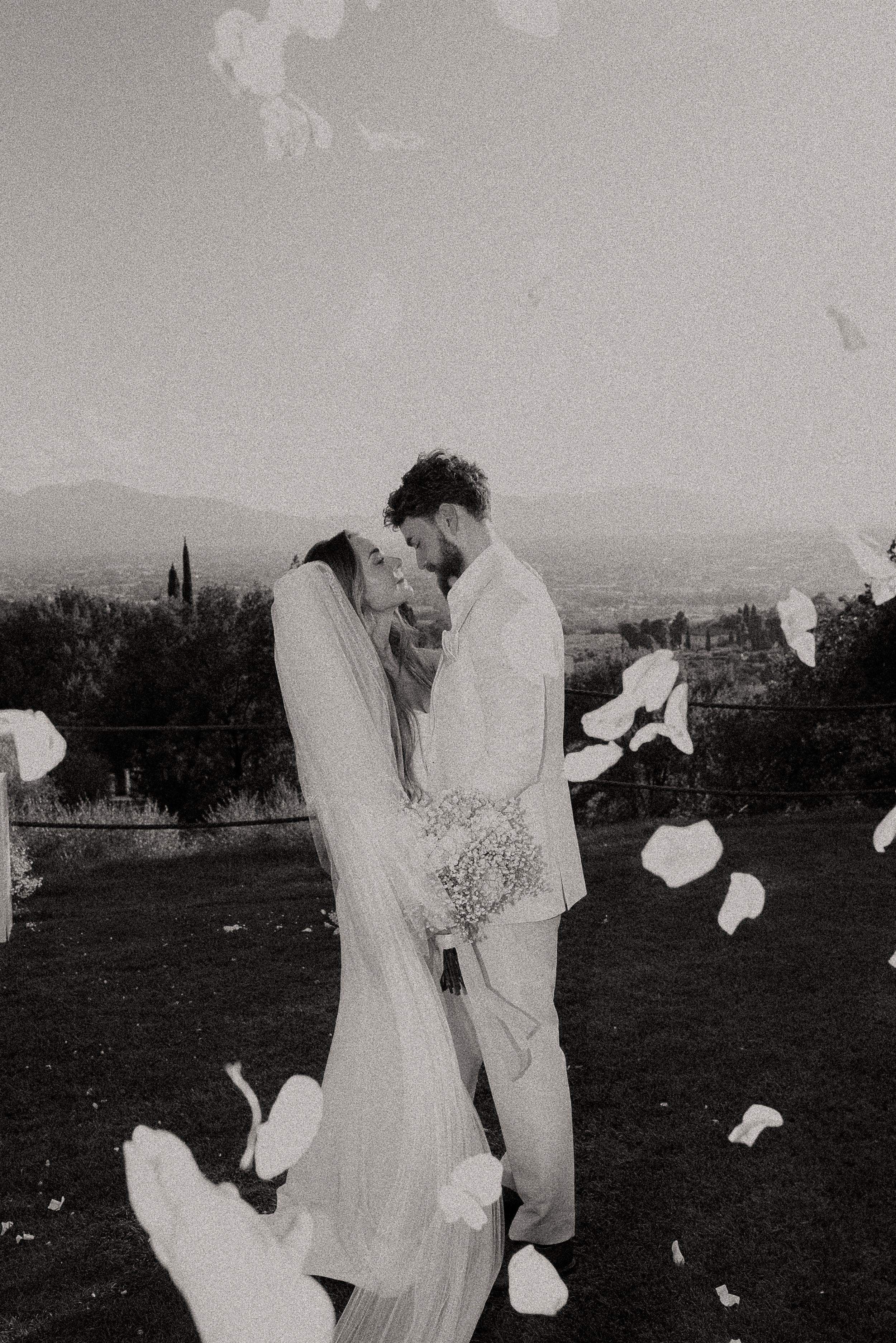 A romantic black and white wedding moment with flower petals floating in the air against a mountain landscape.