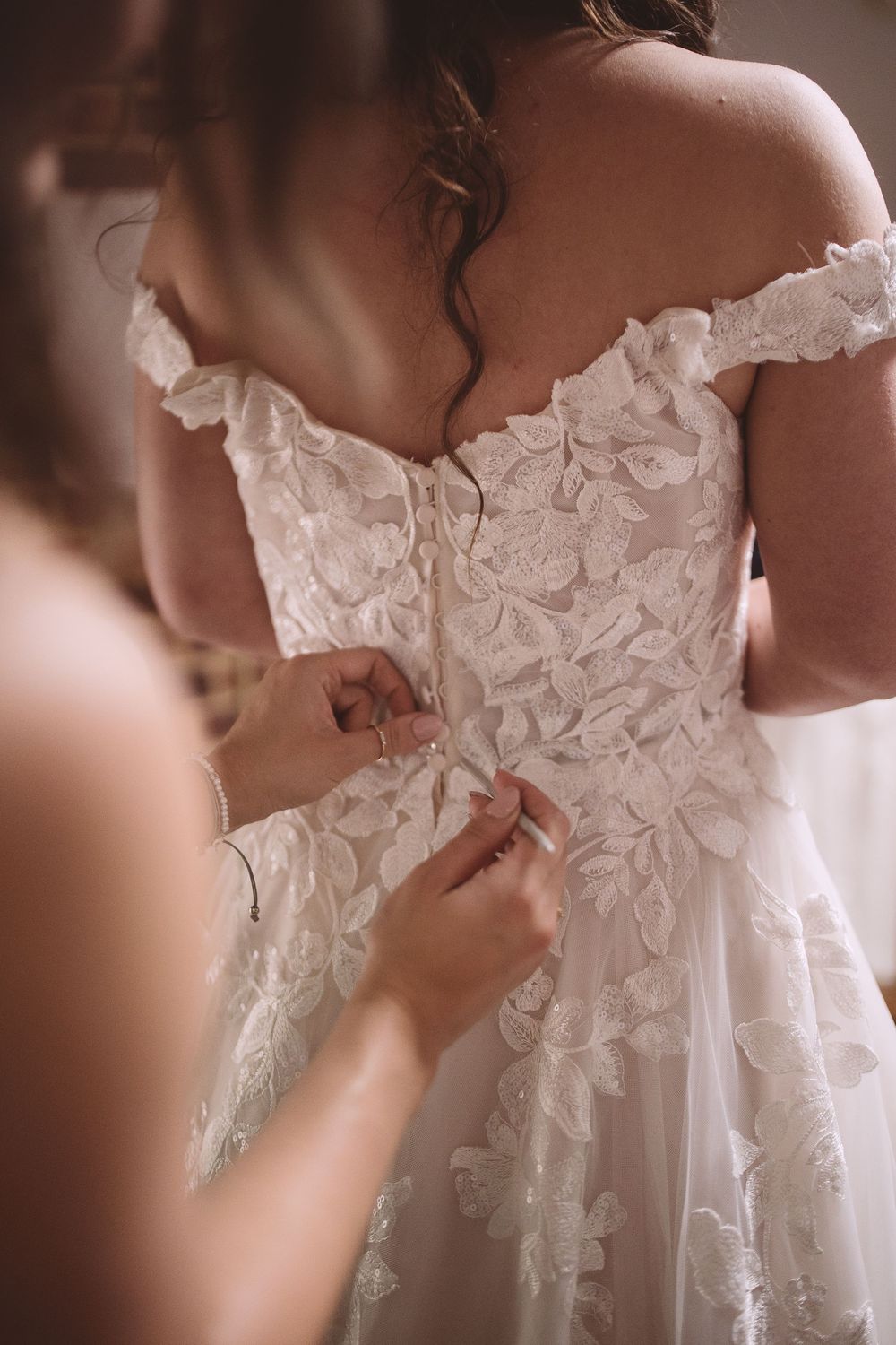 Someone helps fasten the back of an ornate white lace wedding dress with delicate floral details and off-shoulder design.