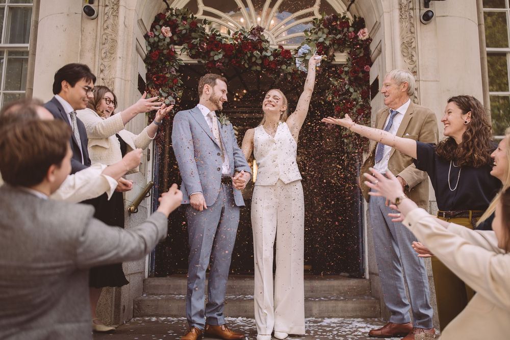 Couple celebrates their wedding day outside historic building entrance with guests showering them in confetti and petals.