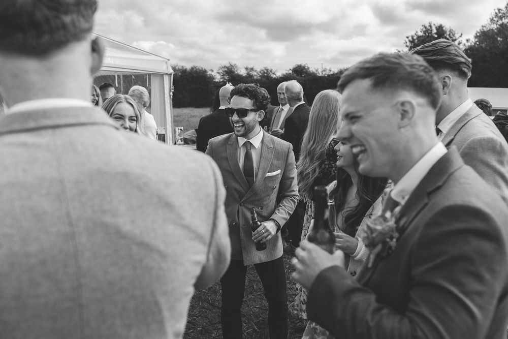 Wedding guests in suits and sunglasses gather outdoors on a cloudy day, captured in black and white photography.