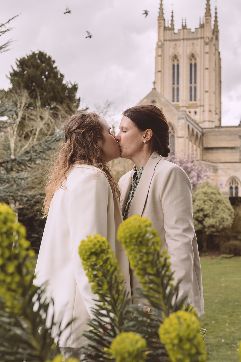 An intimate moment between a same-sex couple standing in front a cathedral with yellow flowers in foreground.