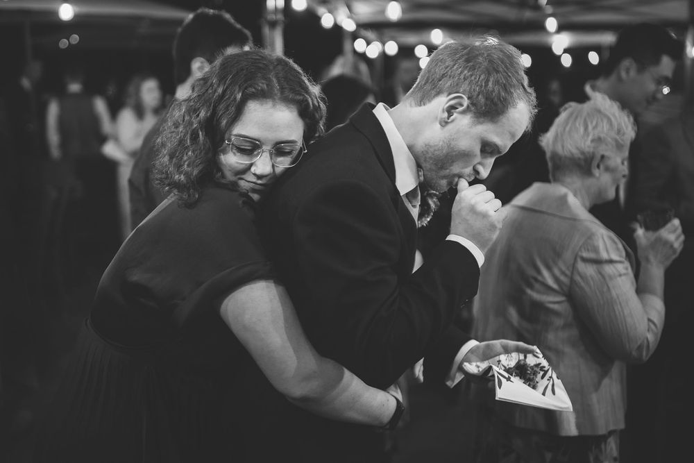 A young woman joyfully embraces a male wedding guest from behind who is entirely focussed on eating cake.
