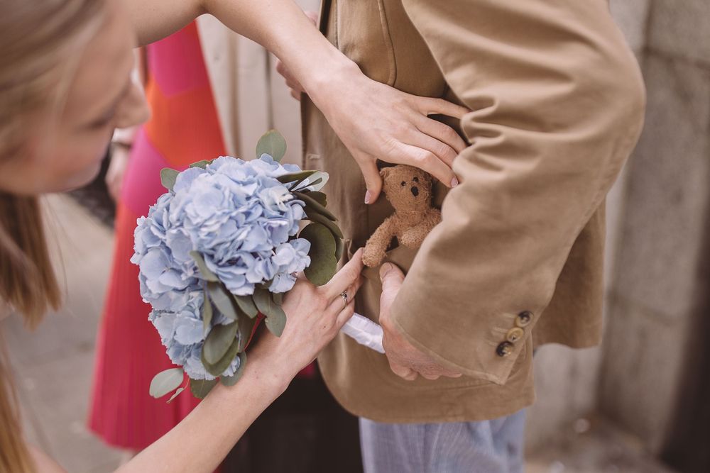 A person holding a delicate blue hydrangea bloom while inserting a teddy into a tan suit jacket.