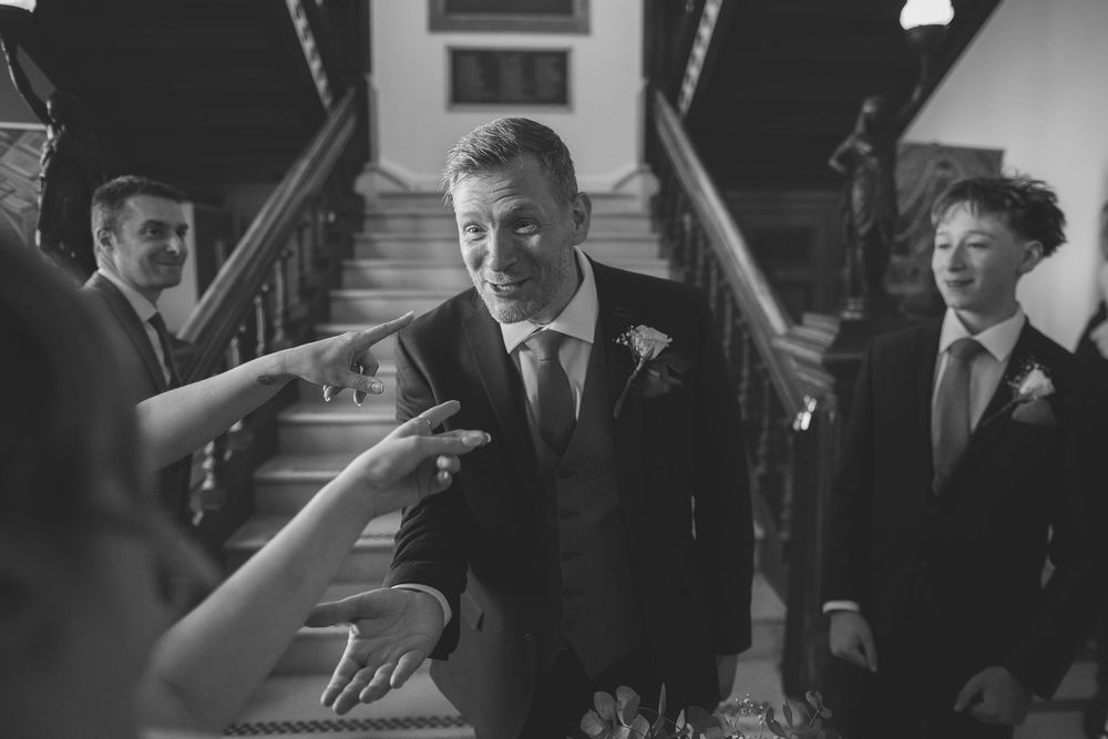 A black and white image captures a candid moment after a wedding ceremony where guests point at the groom.
