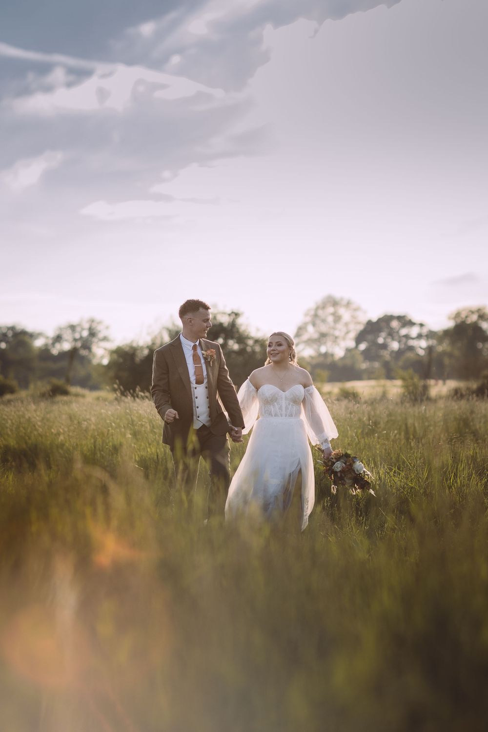 Couple walks hand in hand through tall grass at sunset in a dreamy outdoor suffolk wedding.