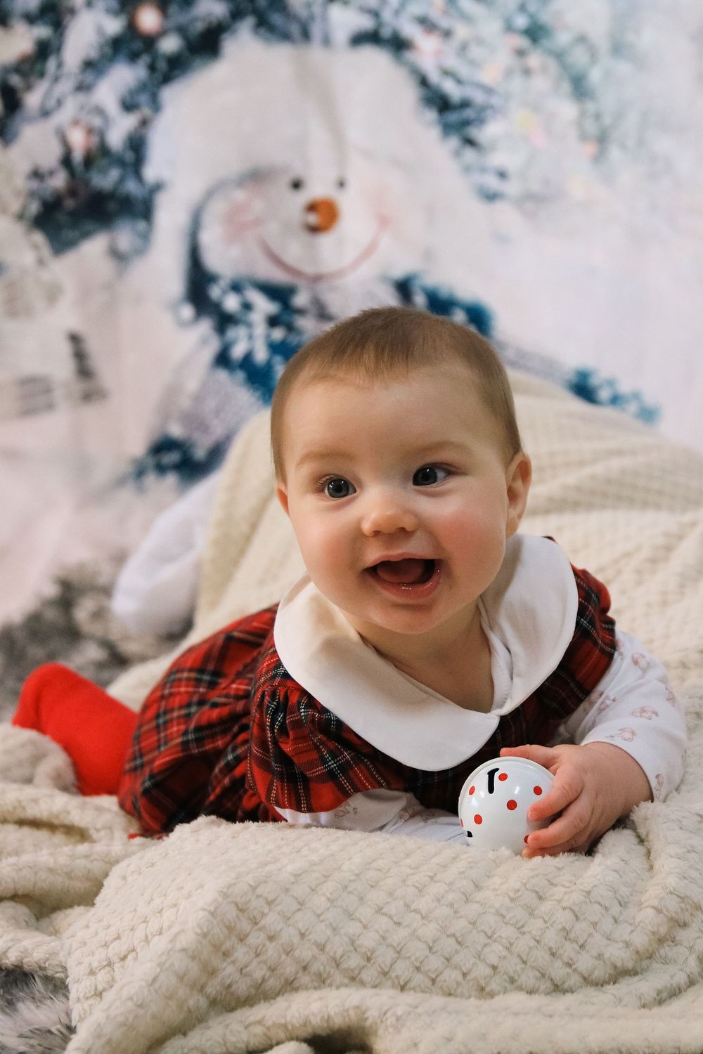 A baby in a red plaid dress lies on a cream textured blanket with winter holiday decorations in the background.