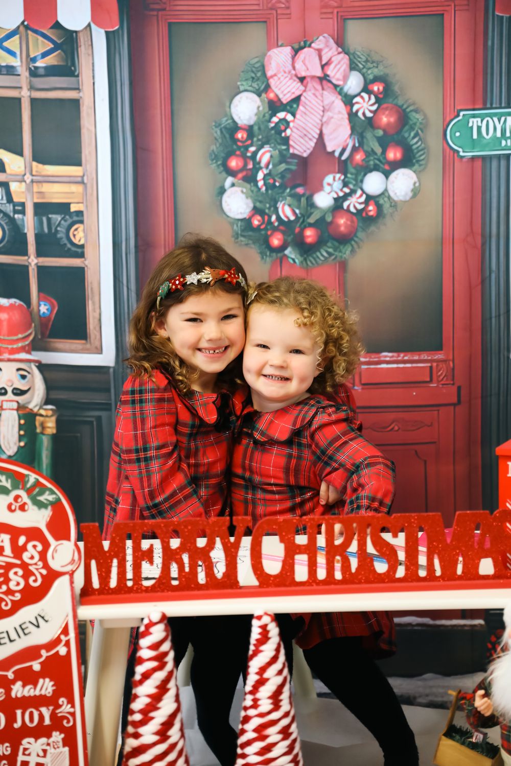 Kids wearing matching red plaid outfits pose in a festive holiday photo studio setting with Christmas wreaths and decorations.