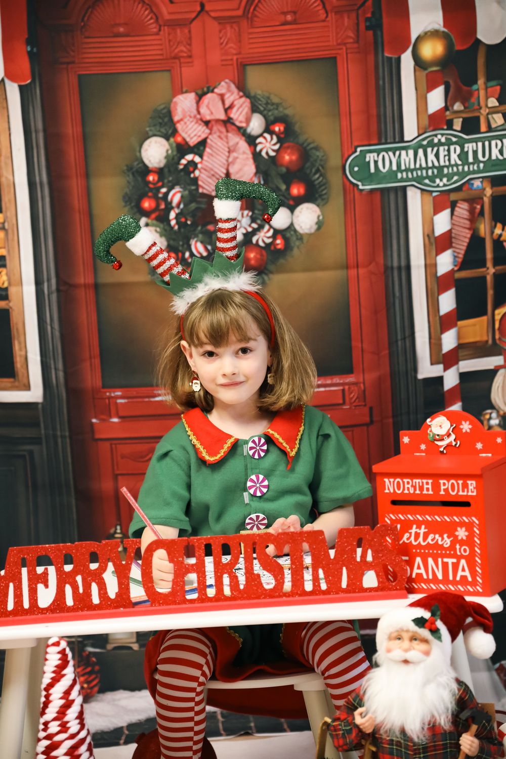 A festive Christmas scene with a green elf outfit against a red door decorated with a wreath and Merry Christmas banner.