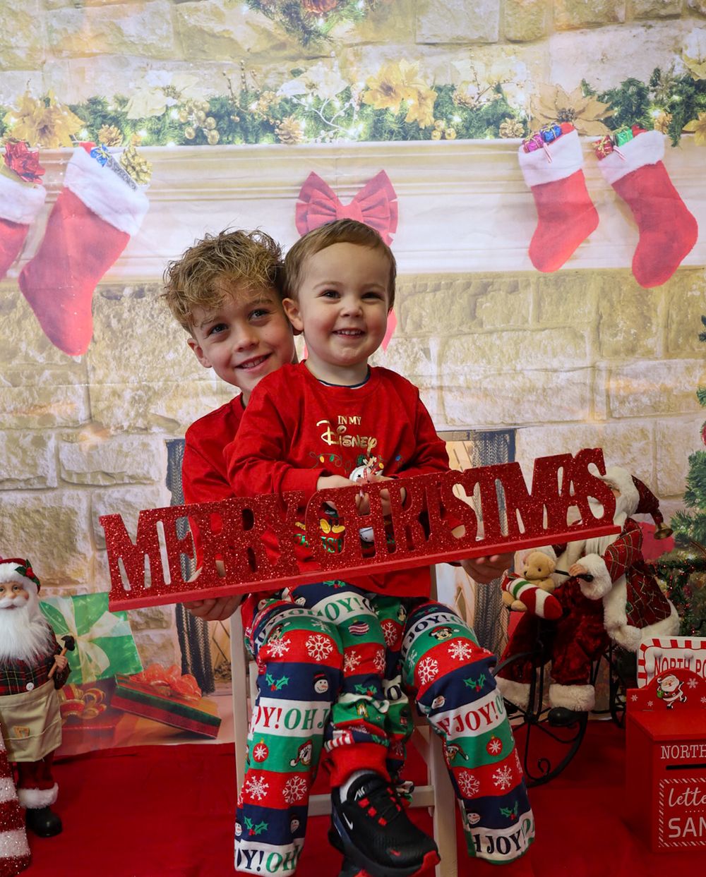 Two children in red holiday attire pose in front of Christmas stockings and decorations on a stone wall.