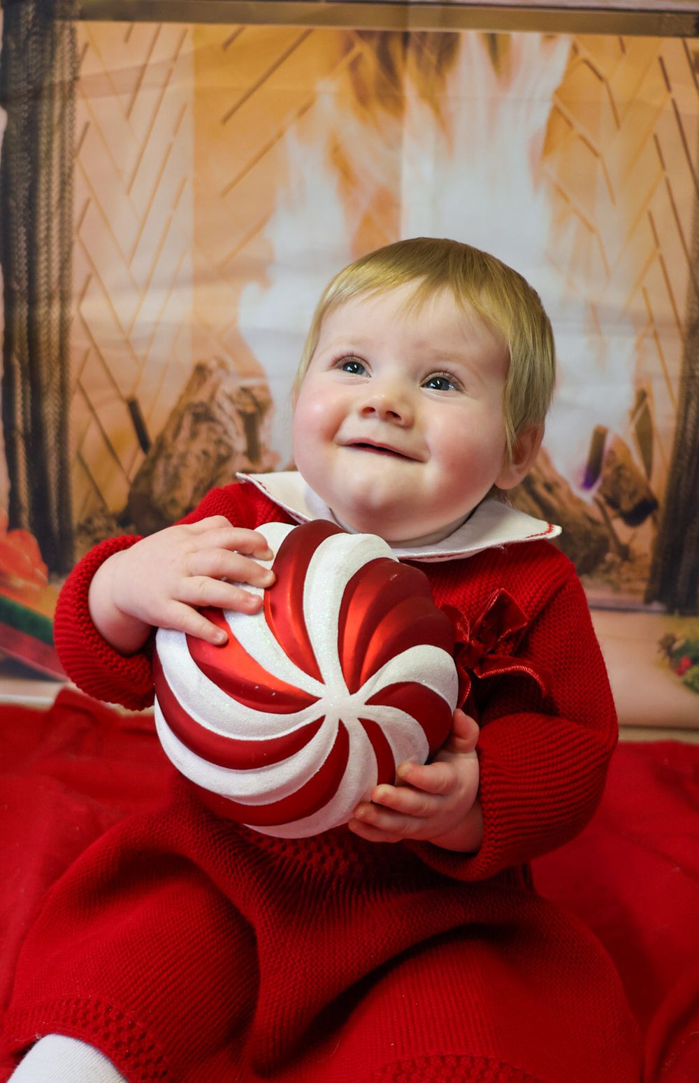 A baby in a red sweater plays with a large red and white striped peppermint candy decoration on a red blanket.