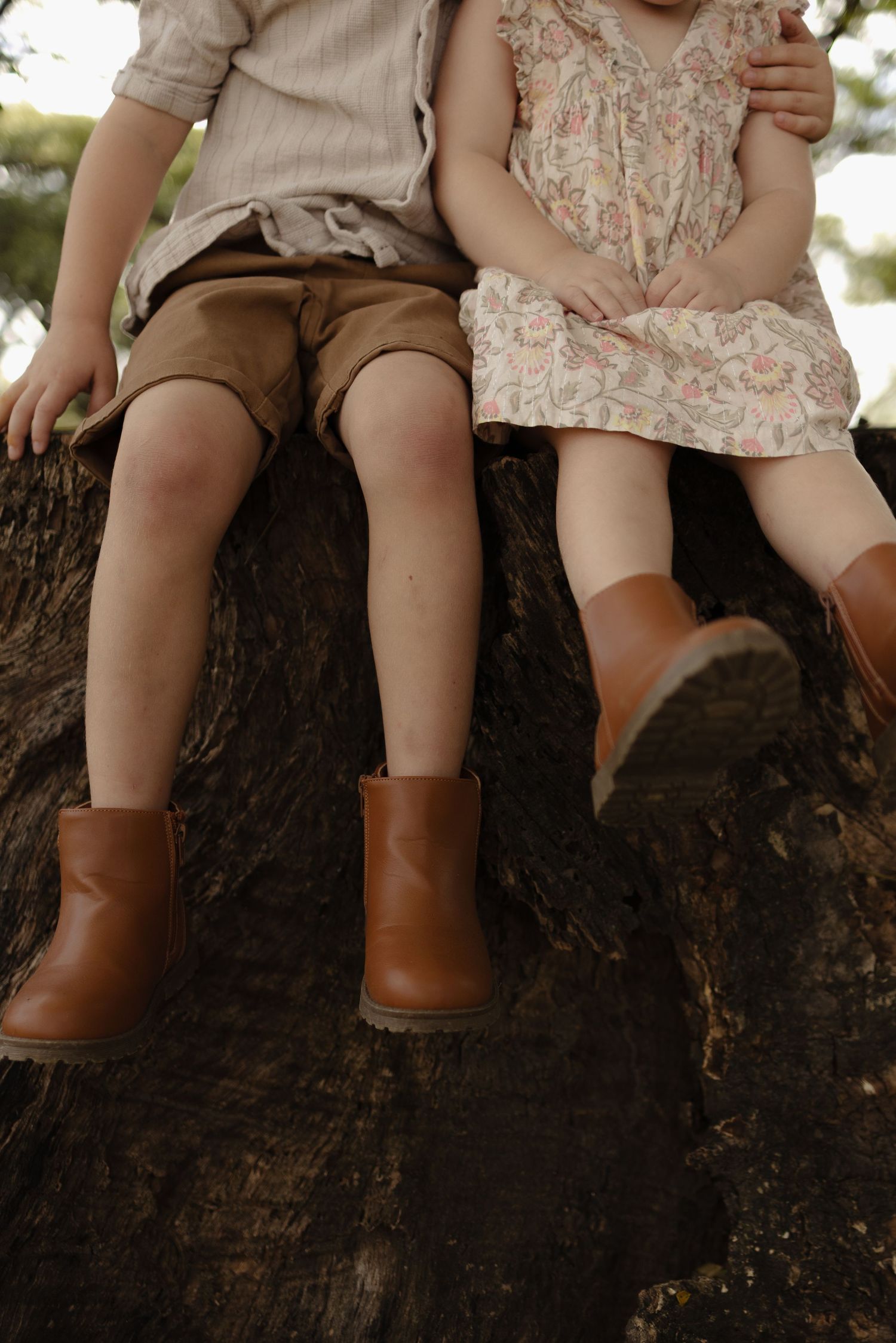 Little feet in brown boots dangle from a wooden log in a natural outdoor setting.