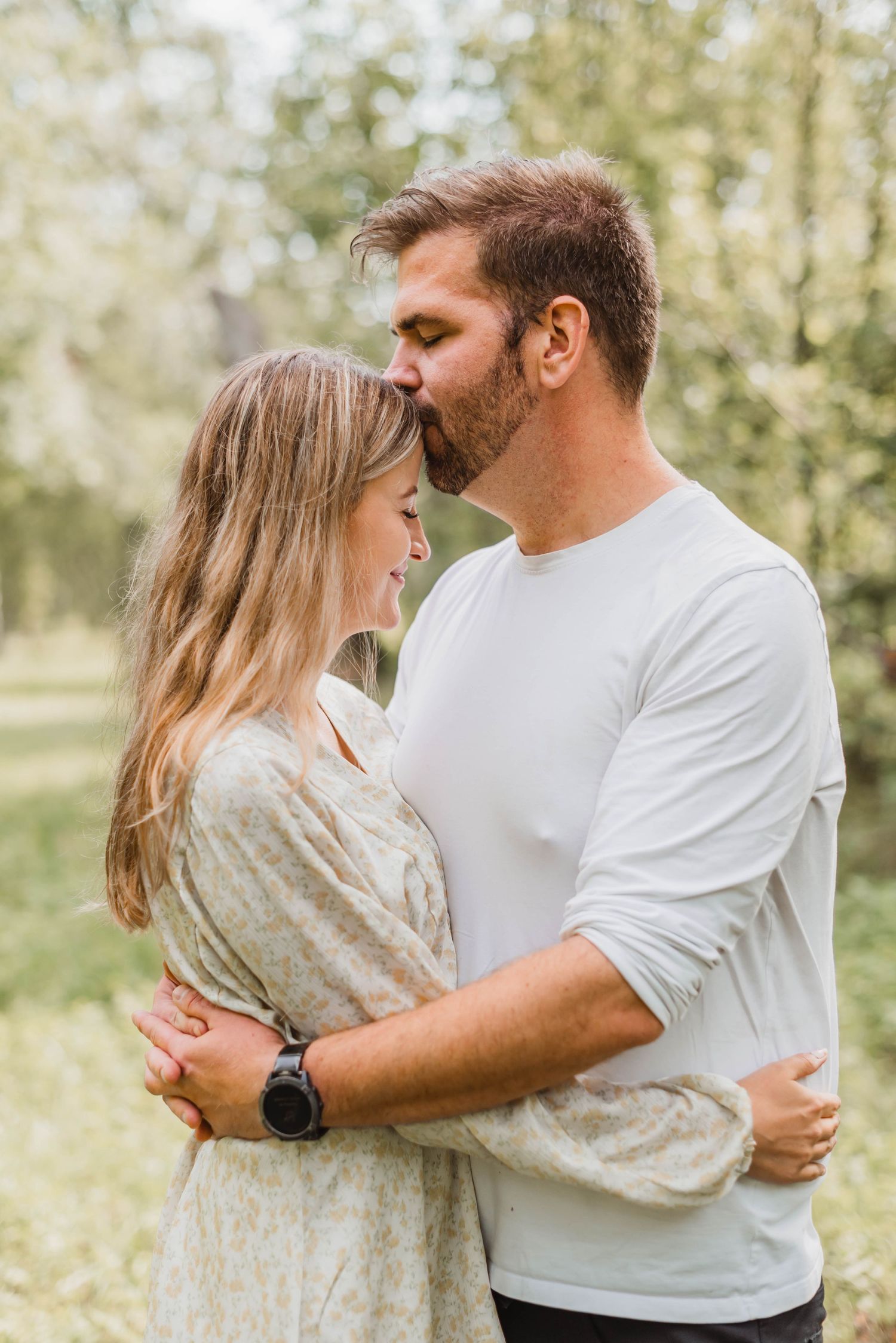 A romantic couple embraces in a soft sunlit park setting wearing coordinated white and beige outfits.