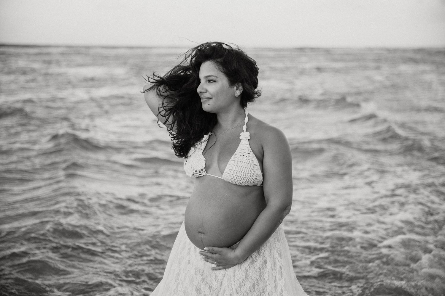 A woman in a white bikini and flowing dress stands by the ocean waves as her hair blows in the wind.