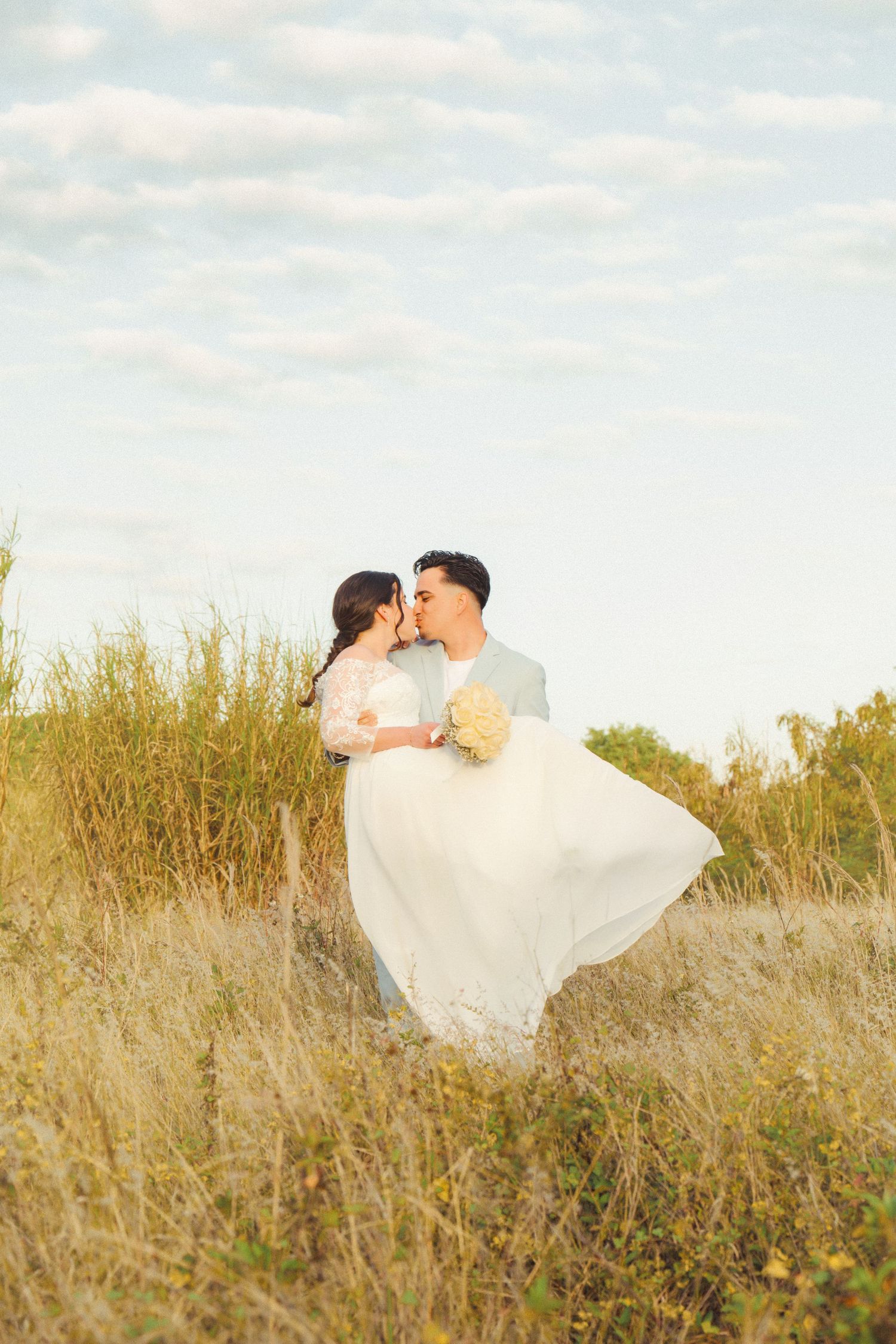 A couple shares a romantic moment in a sunlit field with the bride's dress flowing in the breeze.