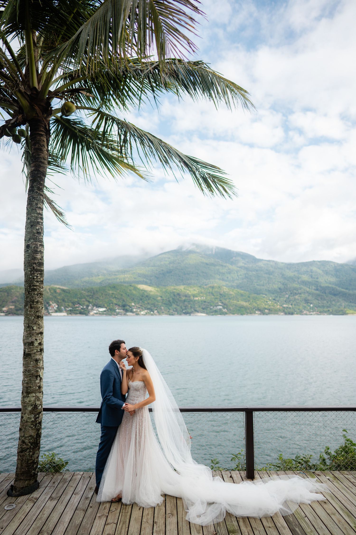 A romantic wedding couple embraces on a wooden deck overlooking a tropical lake with mountains and palm trees in the background.
